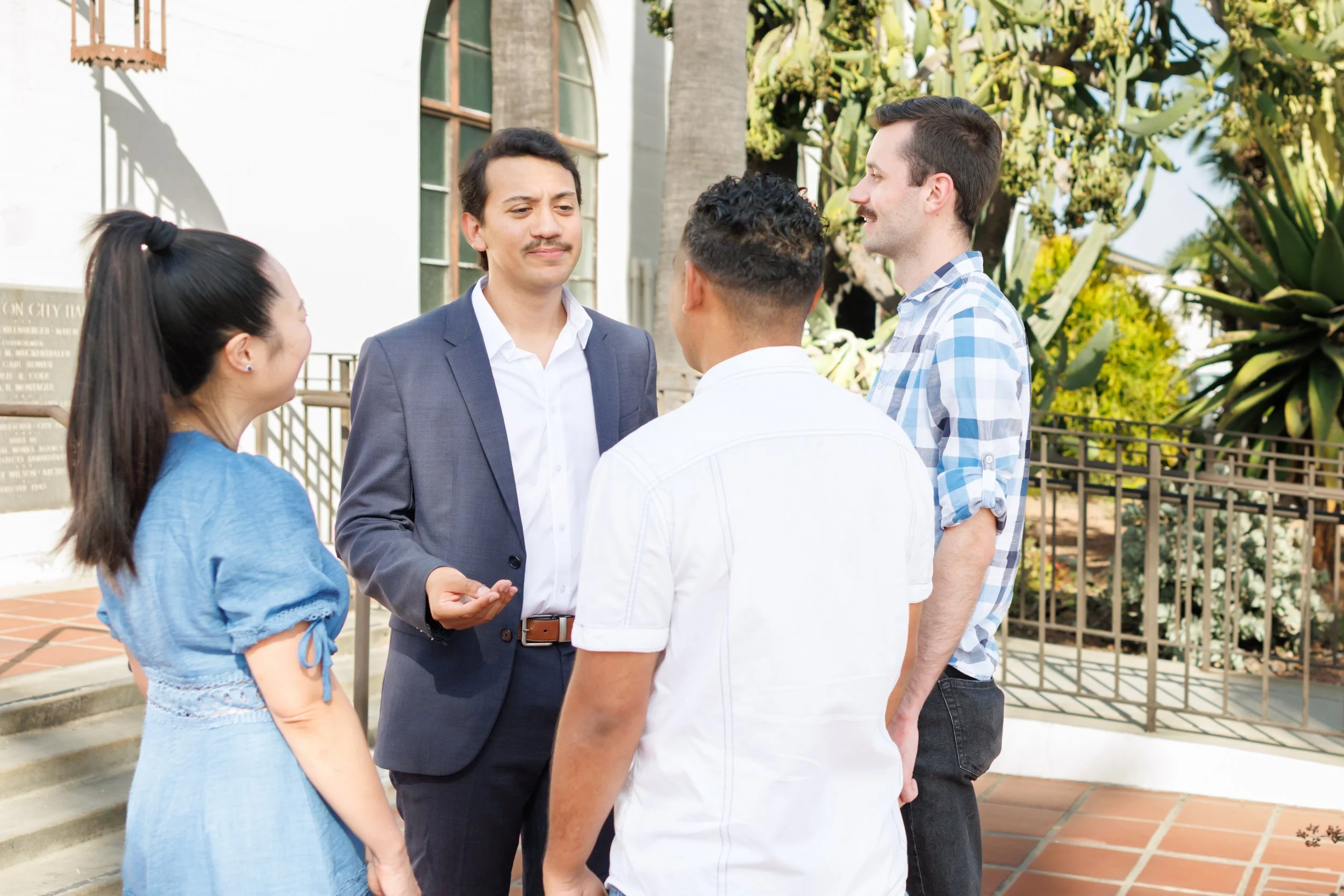 Group of five diverse people standing outside, talking, with a building, a tree, and a metal fence visible in the background.