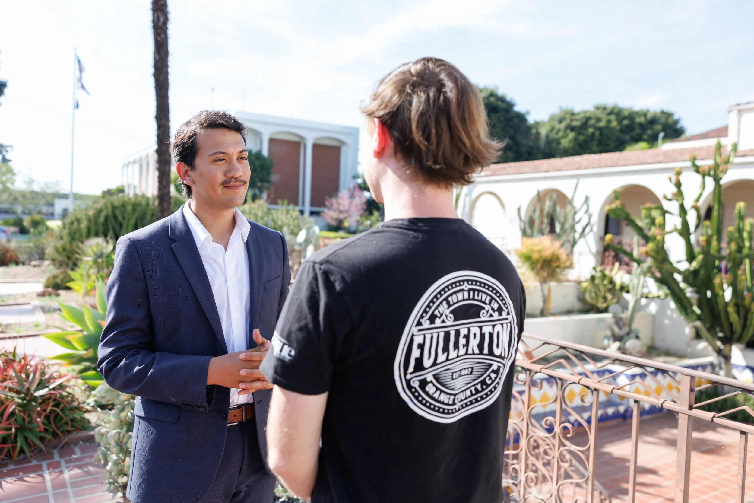 Two men are having a conversation outdoors with greenery and buildings in the background. One is dressed in a navy suit and white shirt, the other in a black T-shirt with a circular white graphic design on the back.
