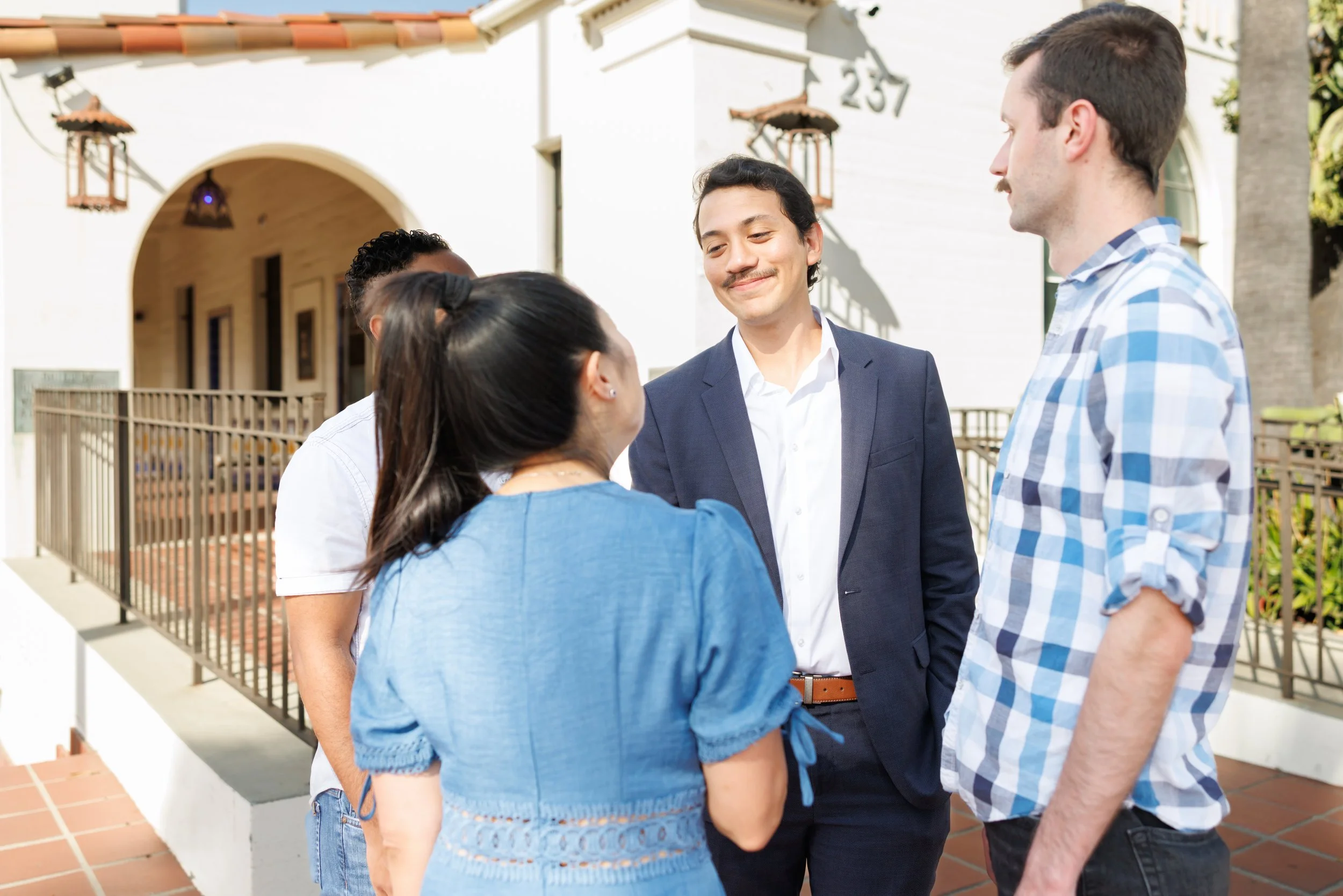 A group of five friends standing together outside a house, engaged in conversation and smiling. They are dressed casually and professionally, indicating a friendly gathering or social event.