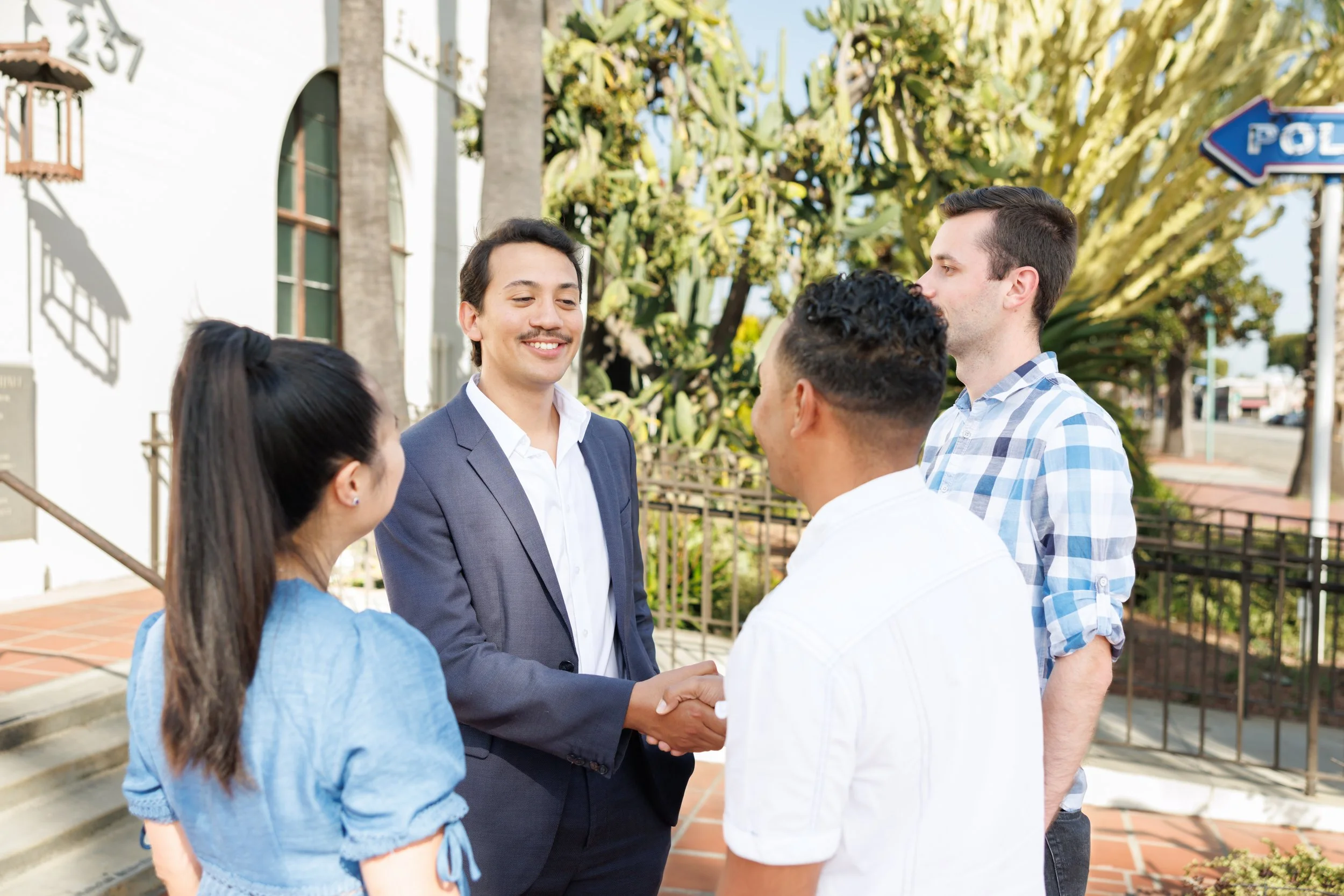 A man in a dark suit and white shirt shaking hands with a woman in a blue outfit, while other people look on during an outdoor event on a sunny day with green trees and buildings in the background.