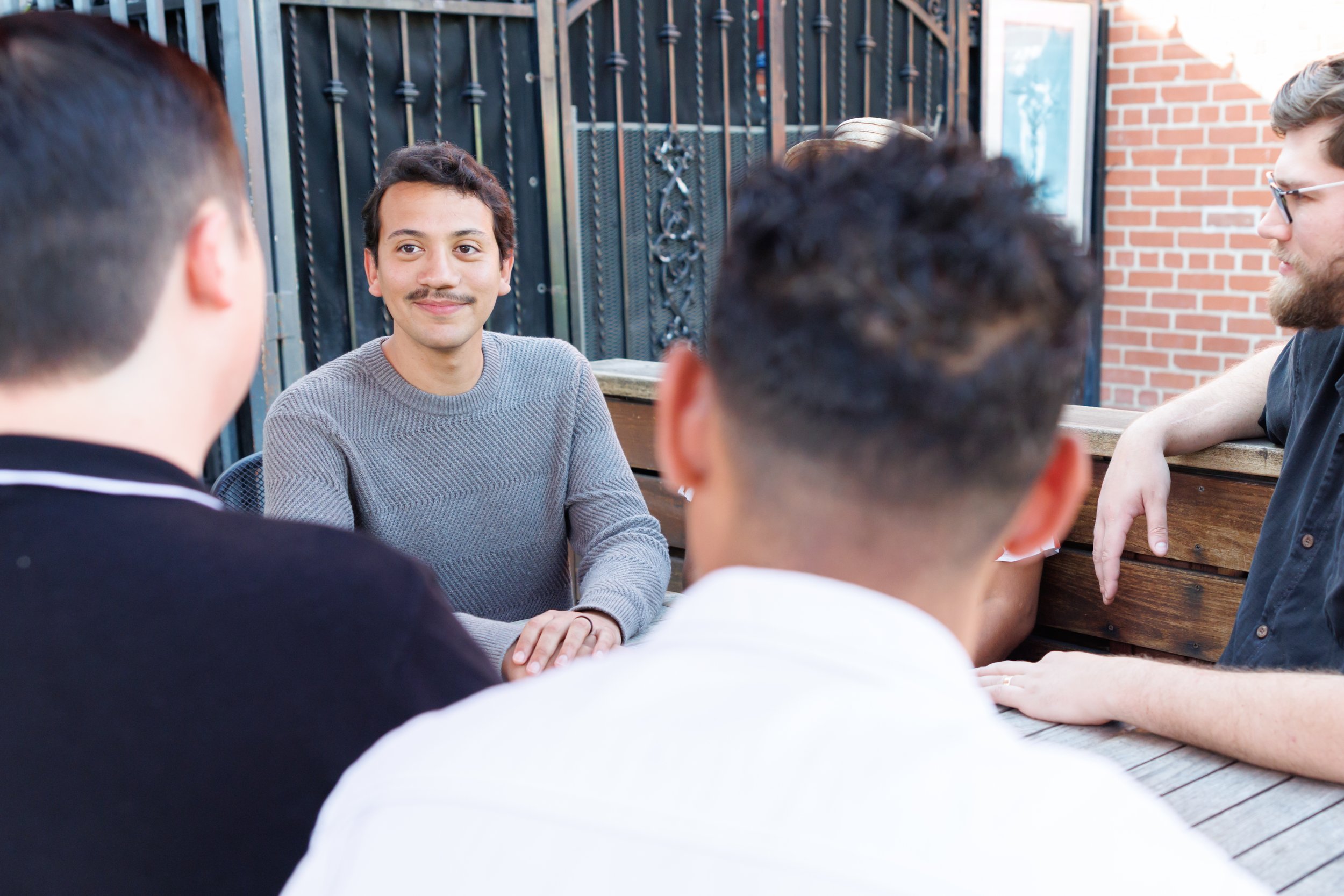 Group of people having a conversation outdoors, with a young man with a mustache smiling in the center, sitting at a wooden table, and other people with diverse hairstyles and glasses facing towards him.