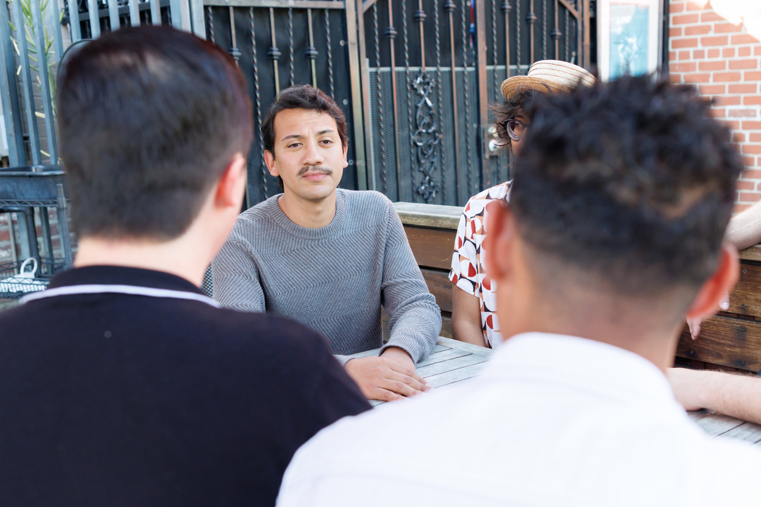 A man with dark hair, a mustache, and wearing a gray sweater is sitting at a table outdoors, listening to three people with their backs to the camera, during daytime. One of the three has curly hair and is wearing a straw hat, and another has short d
