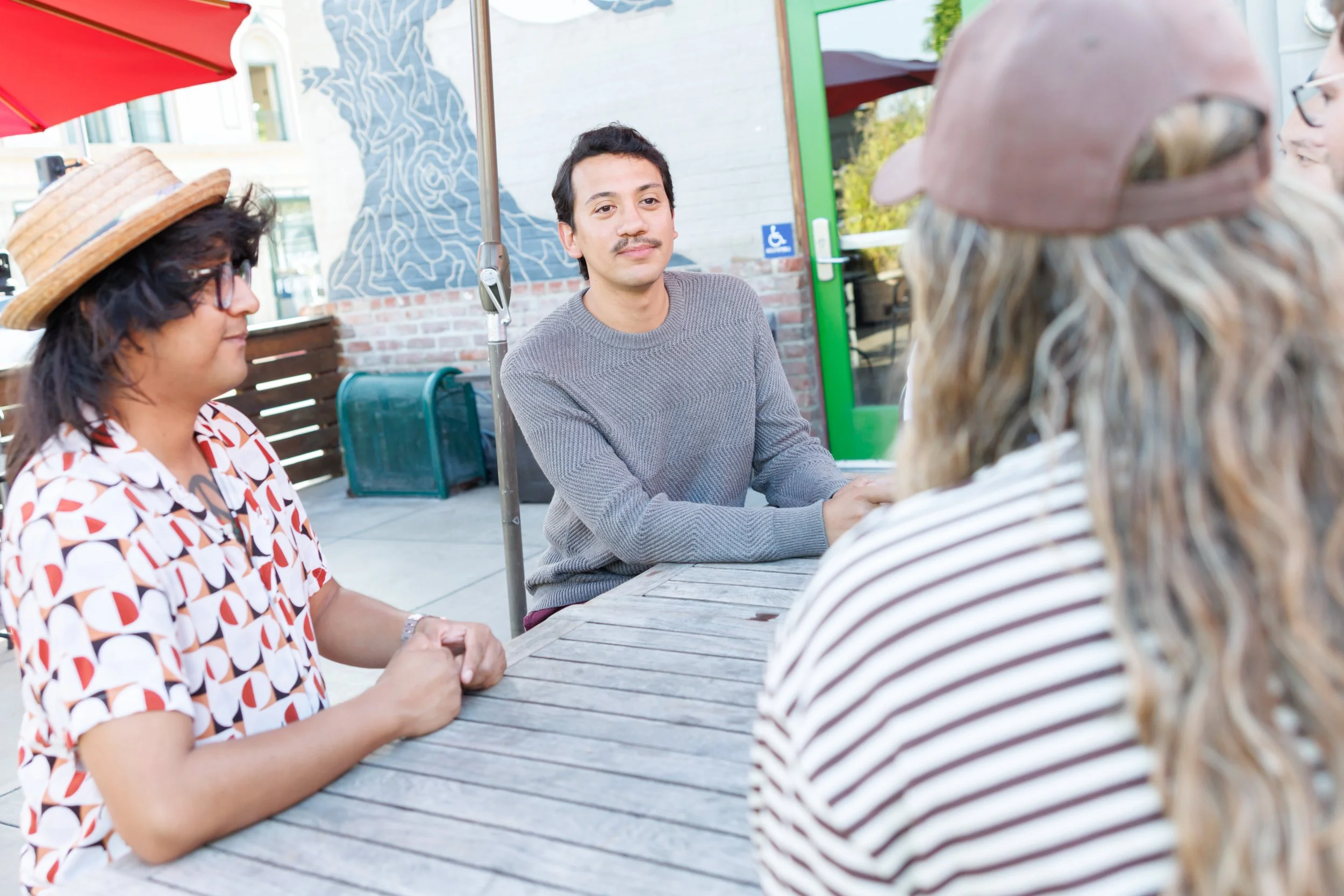 A group of four people sitting at an outdoor wooden table having a conversation. One woman is wearing a patterned blouse and a straw hat, and is holding hands with someone not completely visible. A man in a gray sweater with a mustache is looking att