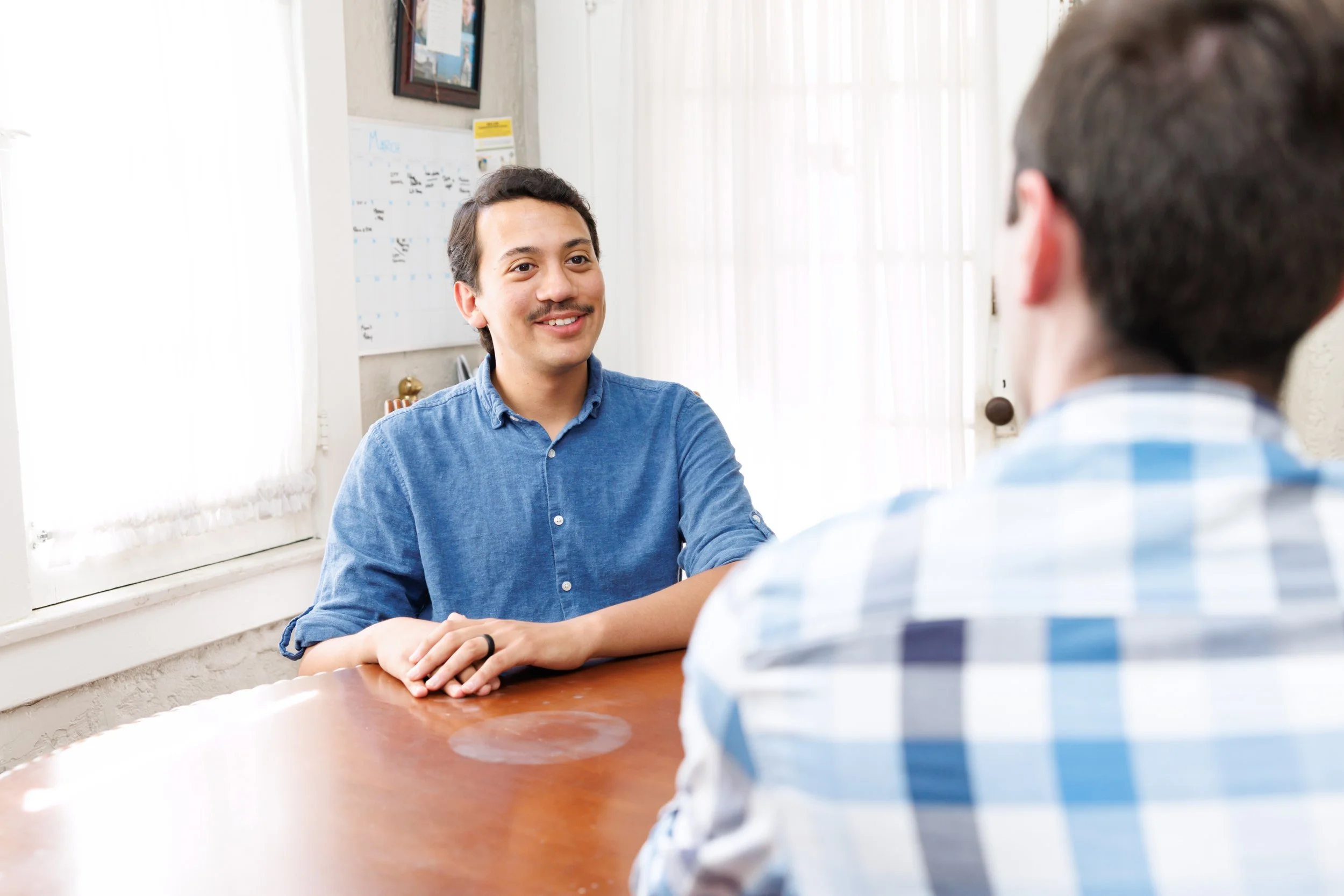 Two men in a discussion in an office, one facing camera with a smile, the other with back to the camera wearing a blue and white checkered shirt.