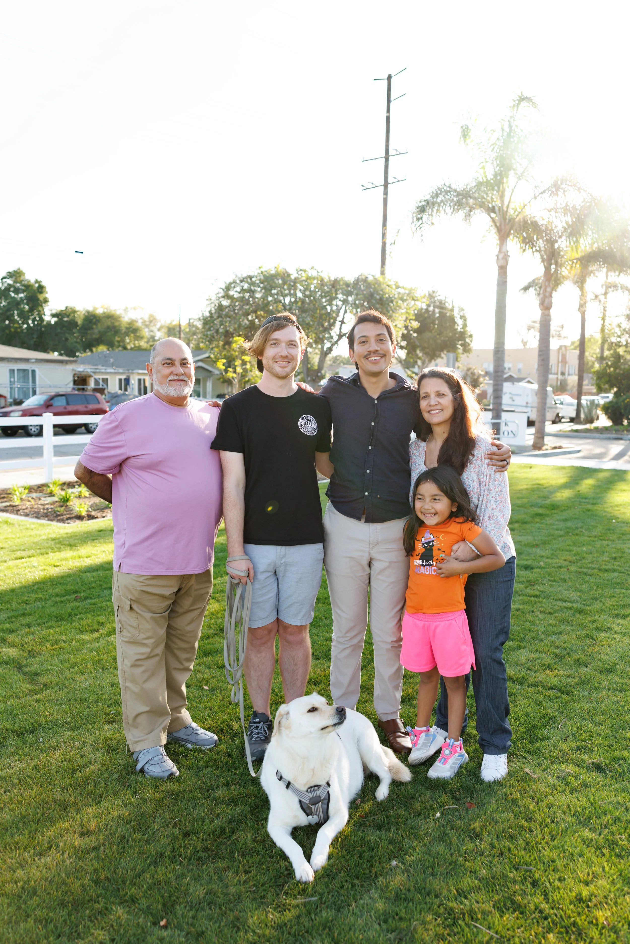 A diverse group of six people, including a woman, a young girl, two young men, an older man, and a woman, stands together on a lawn with sunlight shining behind them, accompanied by a white dog with black spots.
