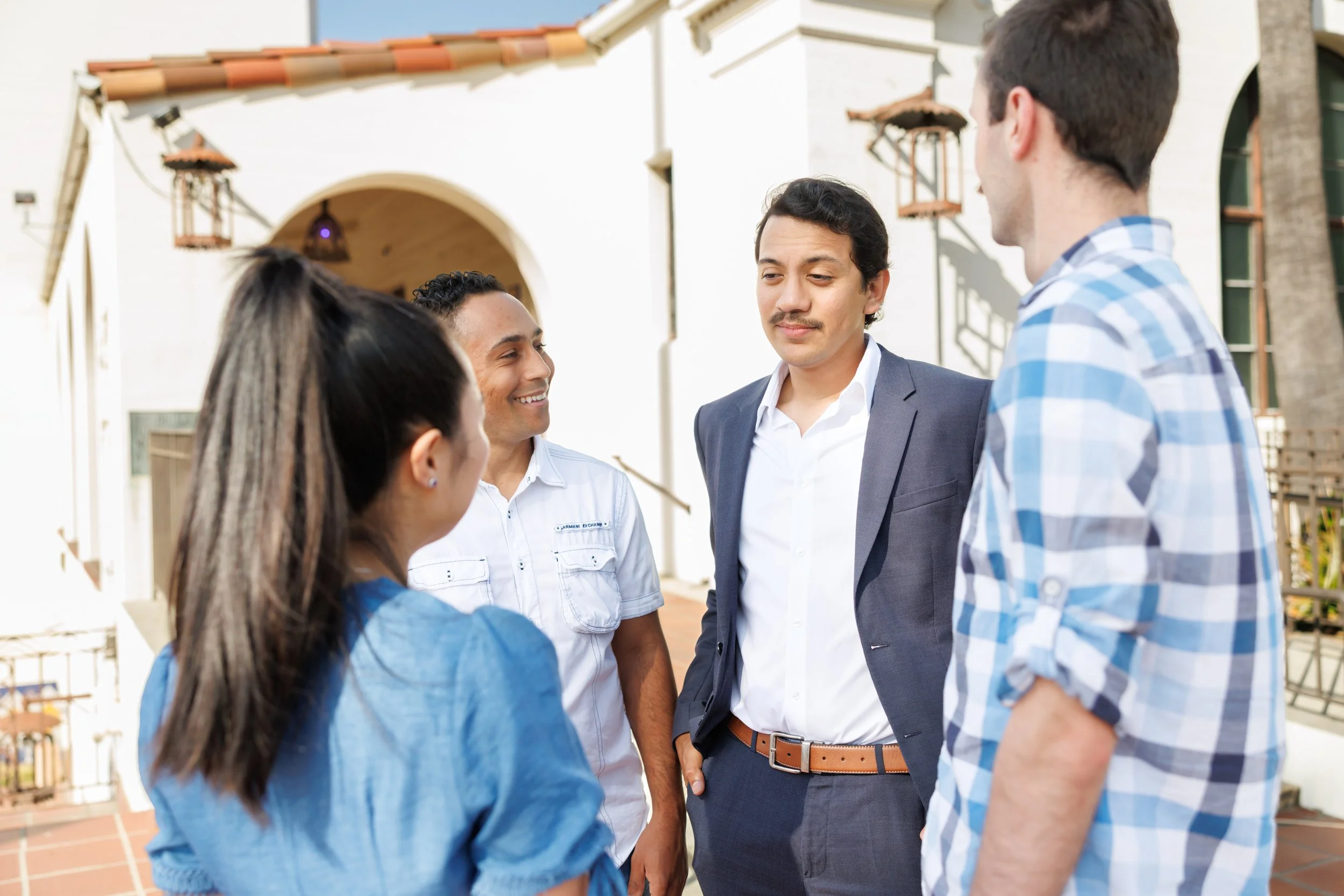Group of four diverse young adults in casual and business attire engaging in a conversation outdoors in front of a white building with a tiled roof.