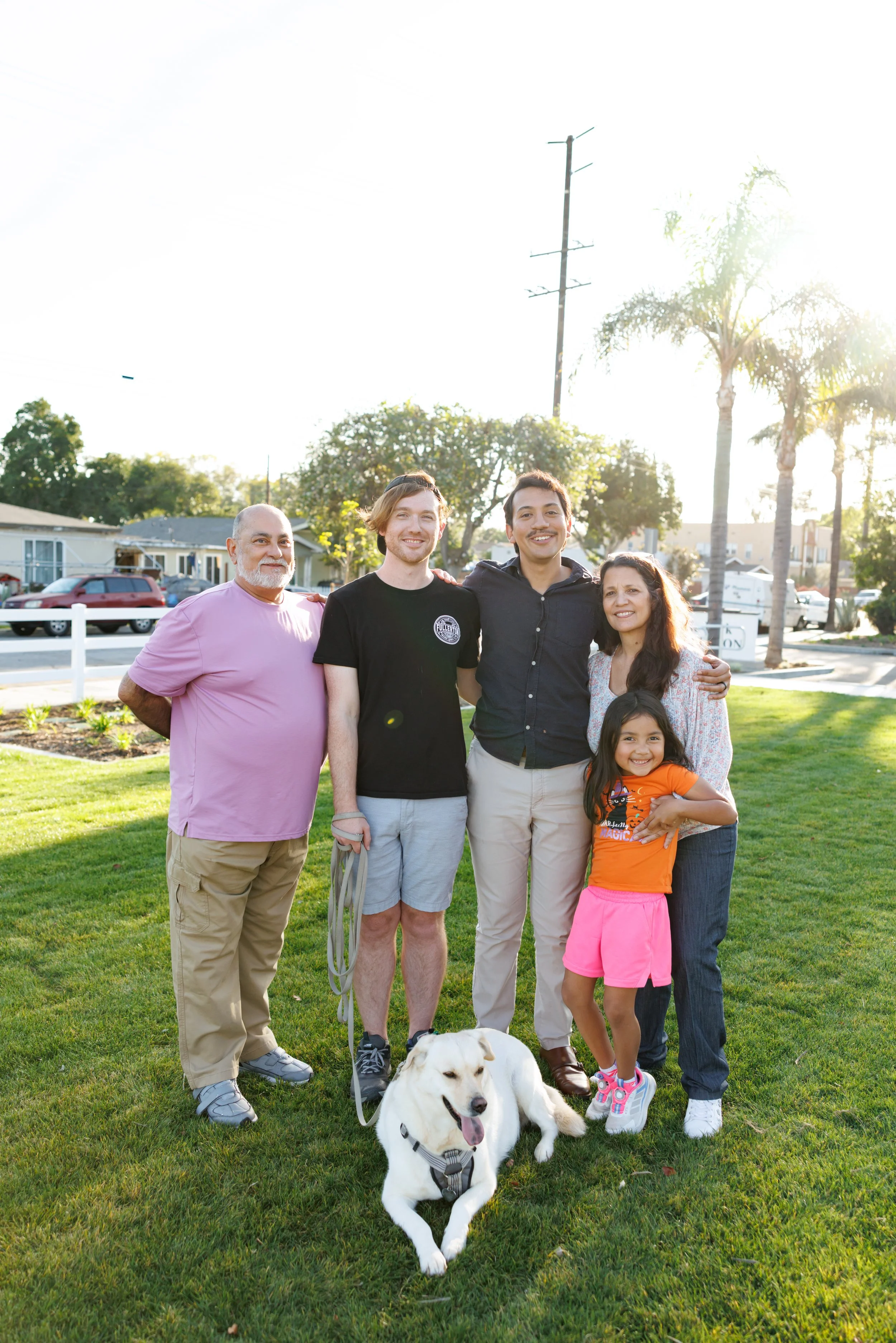 A group of six people, including three men and three women, standing outside on a green lawn, smiling and posing for a photo. There is a young girl in front, and a white dog sitting on the grass, with palm trees, houses, and cars in the background un