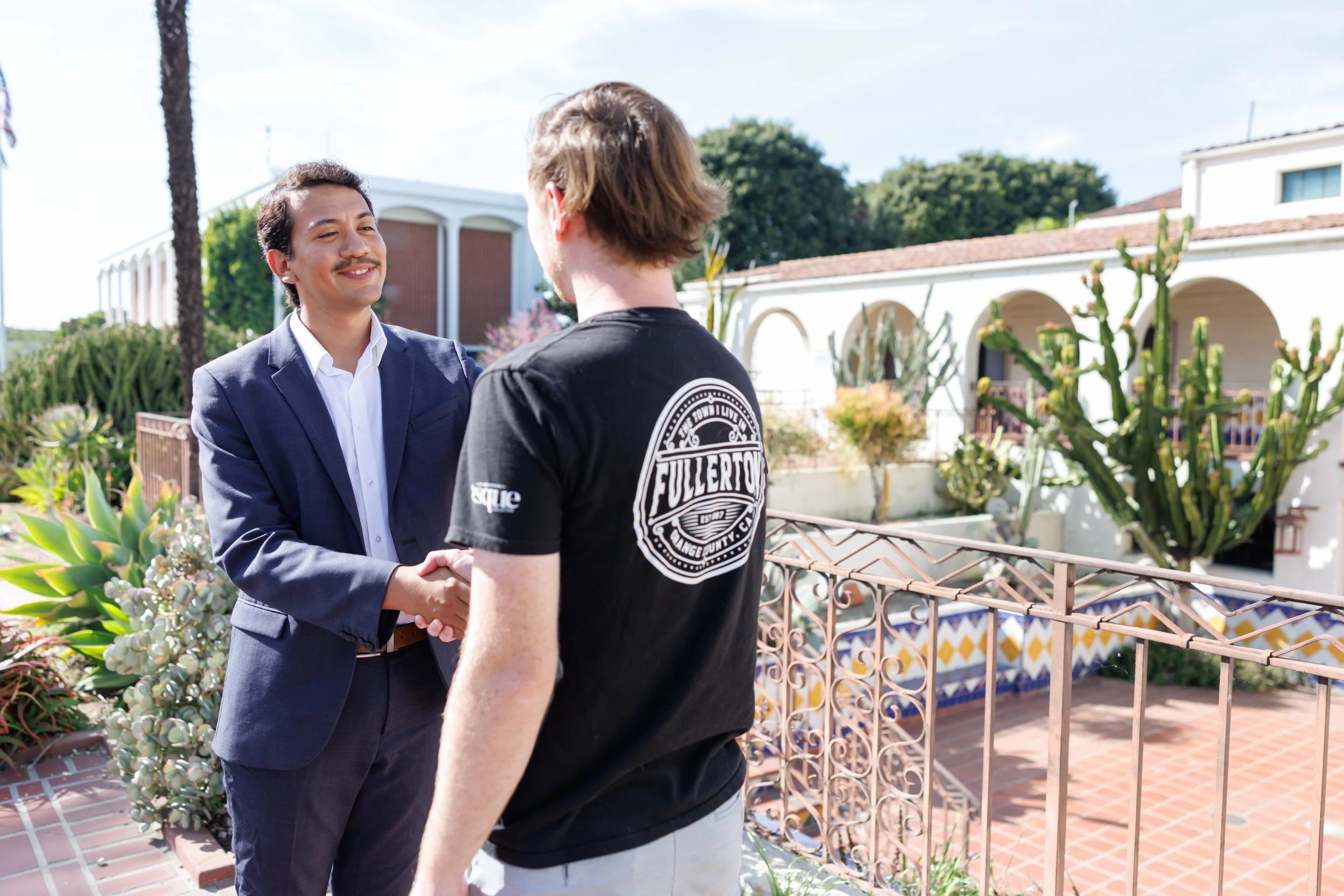 Two men shaking hands outdoors, with a decorative metal railing and potted plants in the background.