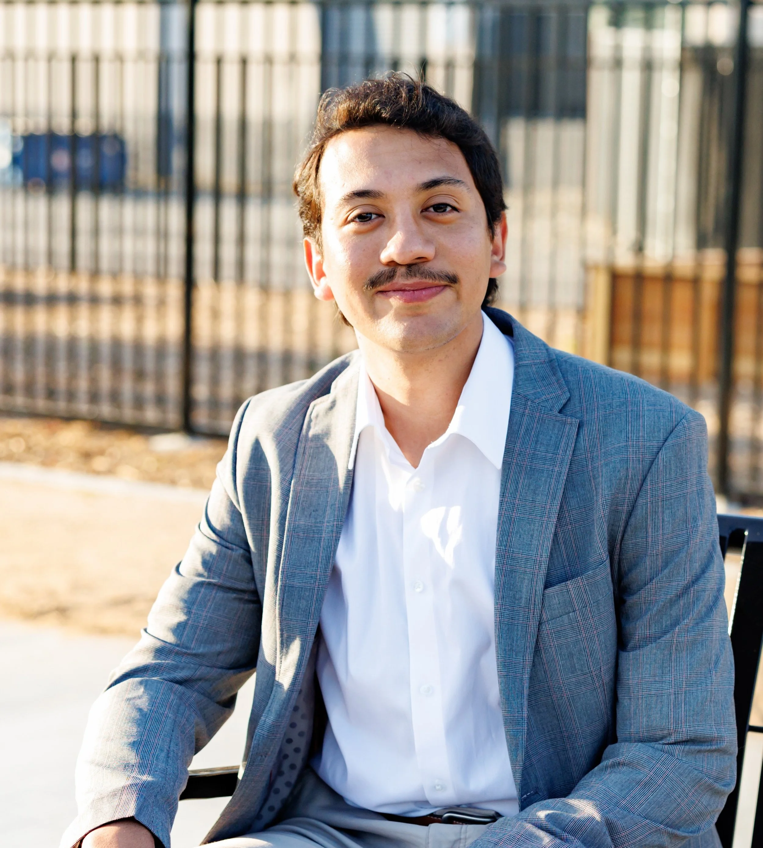 Man in a gray plaid suit and white shirt sitting outdoors in front of a black metal fence, smiling.