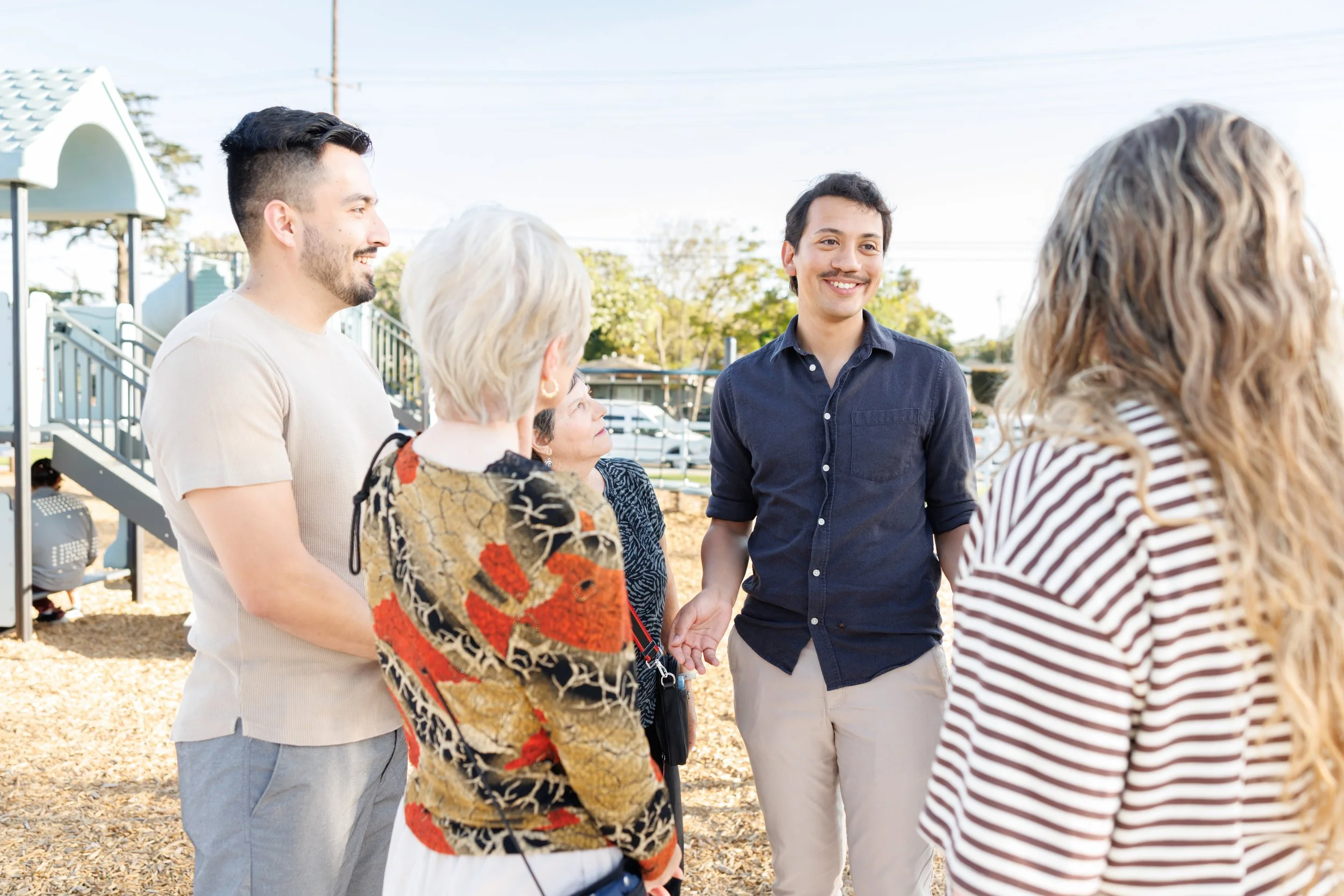 A group of five diverse people, three women and two men, standing outdoors in a circle, talking and smiling on a sunny day. In the background, there's a playground structure and trees.