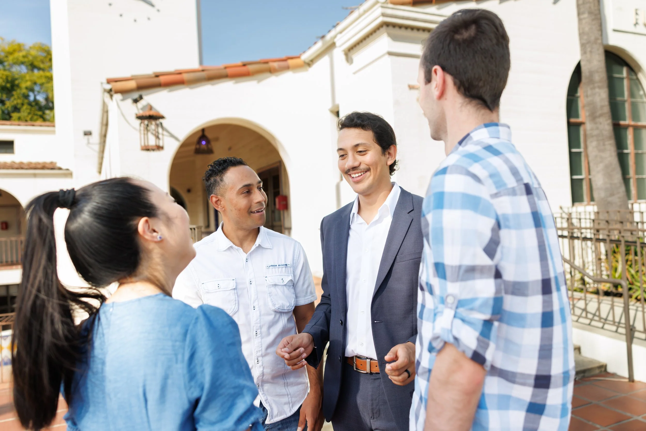 A group of five people standing outside, engaging in a conversation. One man is shaking hands with another man, while the others are watching and smiling. The background shows a white building with arched entryways and a tile roof.