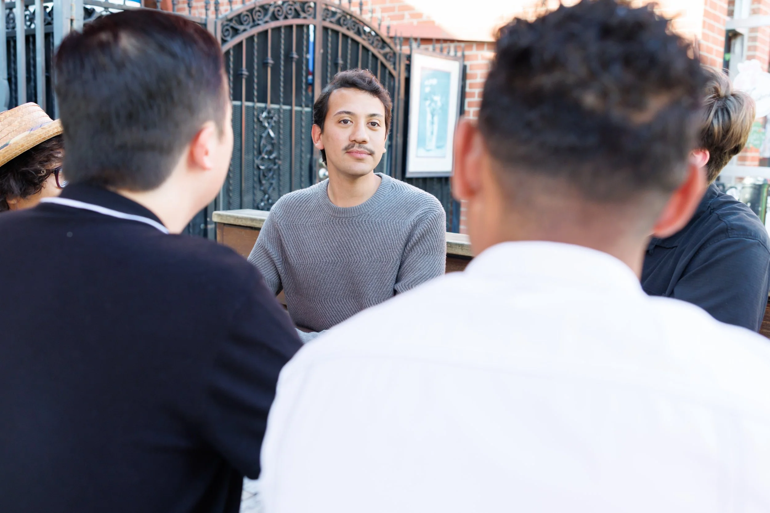 A young man with dark hair and a mustache wearing a gray sweater sitting outdoors at a table with three other people. The image is taken from behind, focusing on the man and the backs of the others, with a decorative iron fence and brick building in 