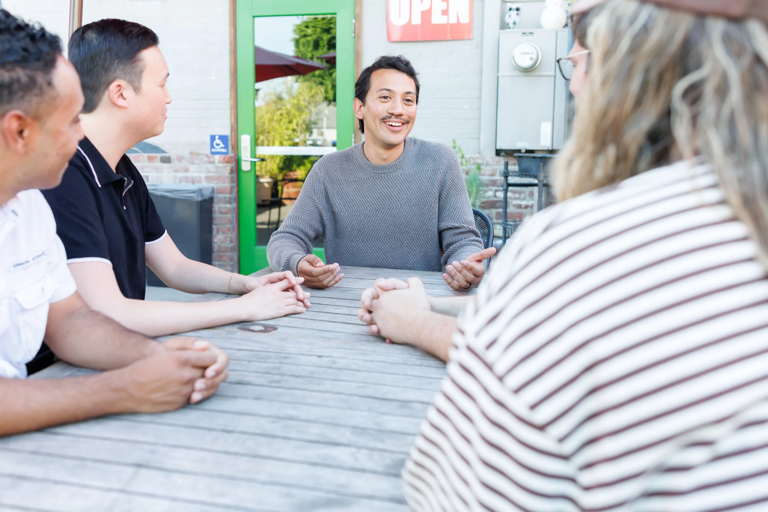 A man speaking to a group of diverse people sitting around an outdoor table, with a green door and a red "Open" sign in the background.
