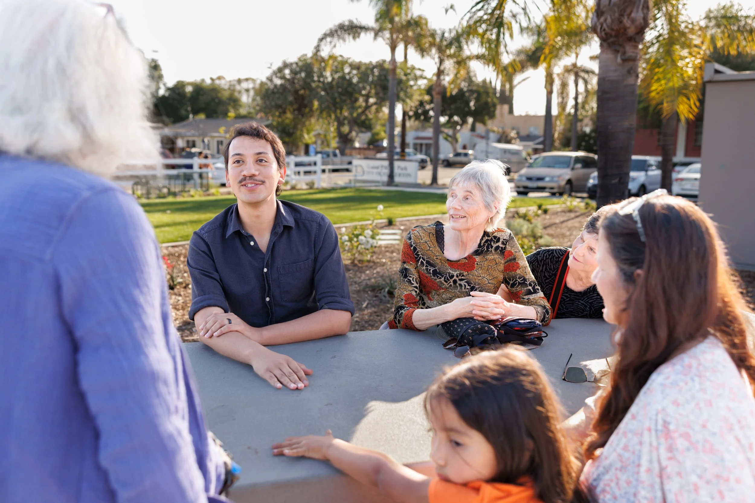Group of people, including a young man with a mustache and elderly women, gathered outdoors around a table, engaging in conversation during daytime with trees and parked cars in the background.