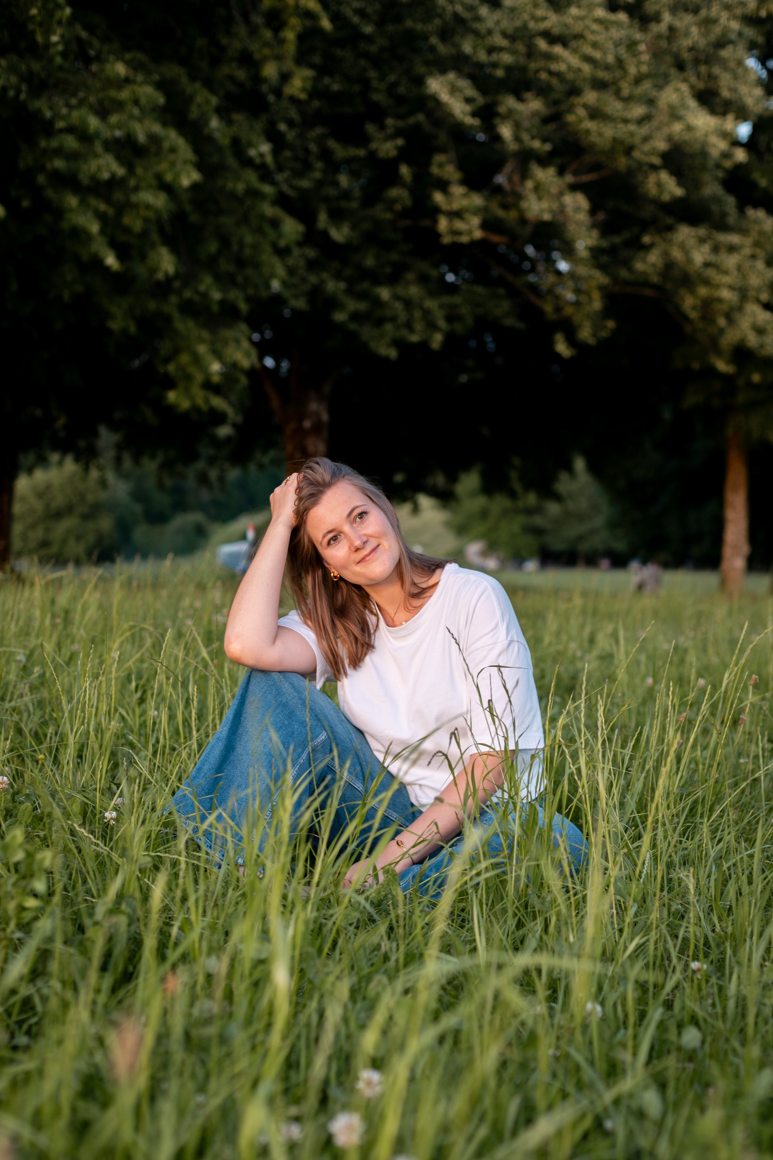 A young woman with long brown hair sitting on green grass in a park, leaning her head on her hand and smiling, with tall trees in the background during late afternoon or early evening.