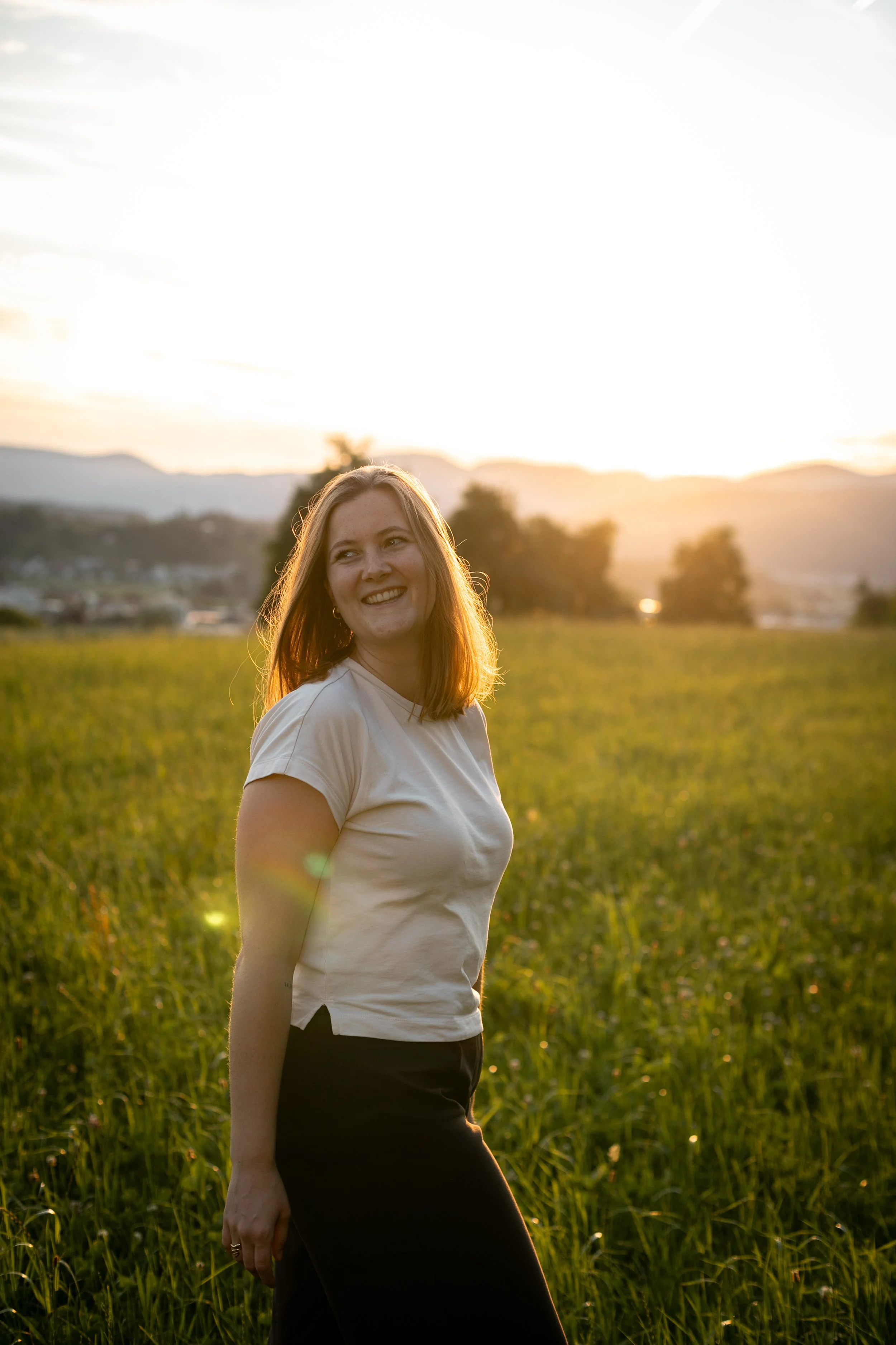 A woman with shoulder-length hair smiling in a field during sunset, with mountains in the background and the sky brightly lit.
