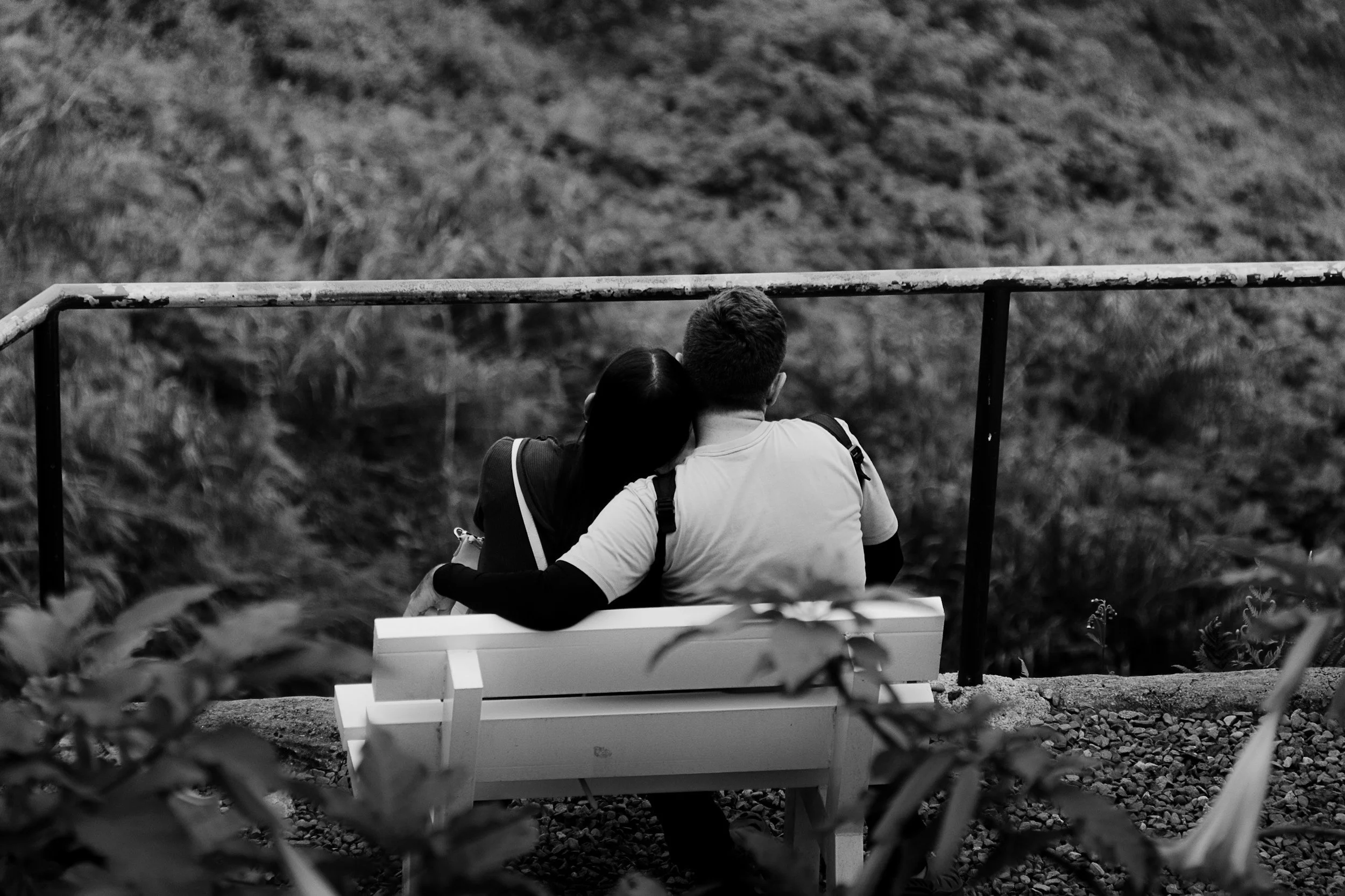 Black and white photo of a couple sitting closely on a park bench, sharing a quiet moment of connection and support in a natural setting.