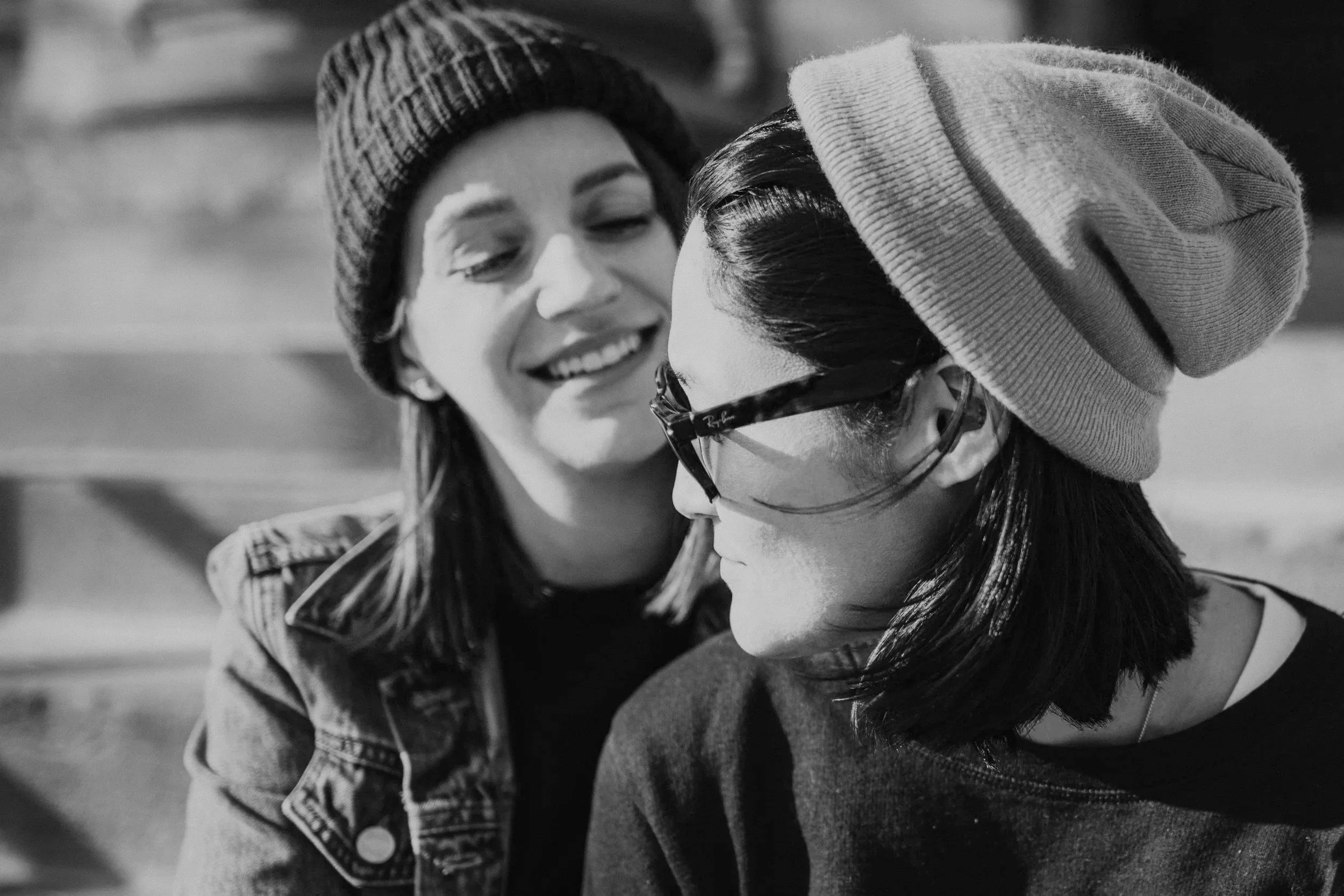 Two young women wearing winter hats and jackets, close-up shot of their faces, smiling and looking at each other in an outdoor setting.