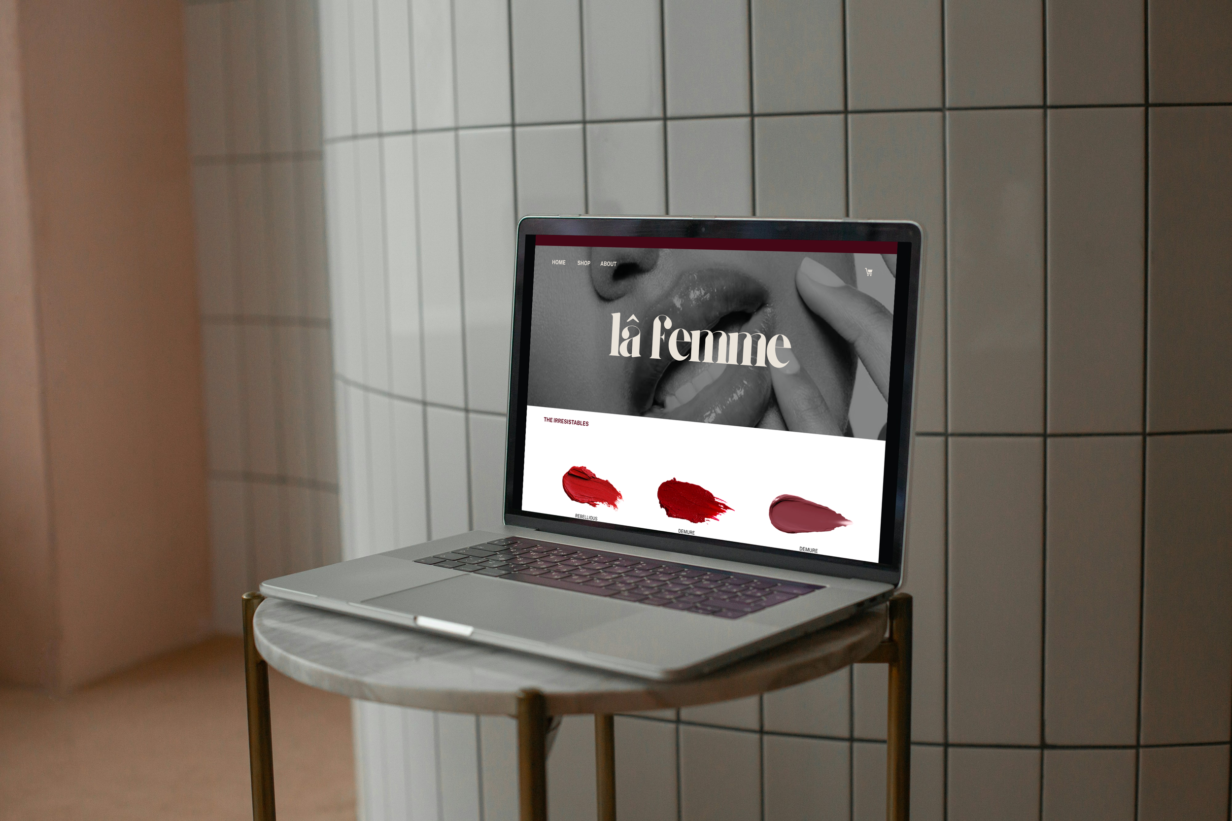 A silver laptop displaying a website titled 'la femme' featuring different shades of lipstick, including 'rebellious' and 'demure,' placed on a small marble table next to a wall with white rectangular tiles.