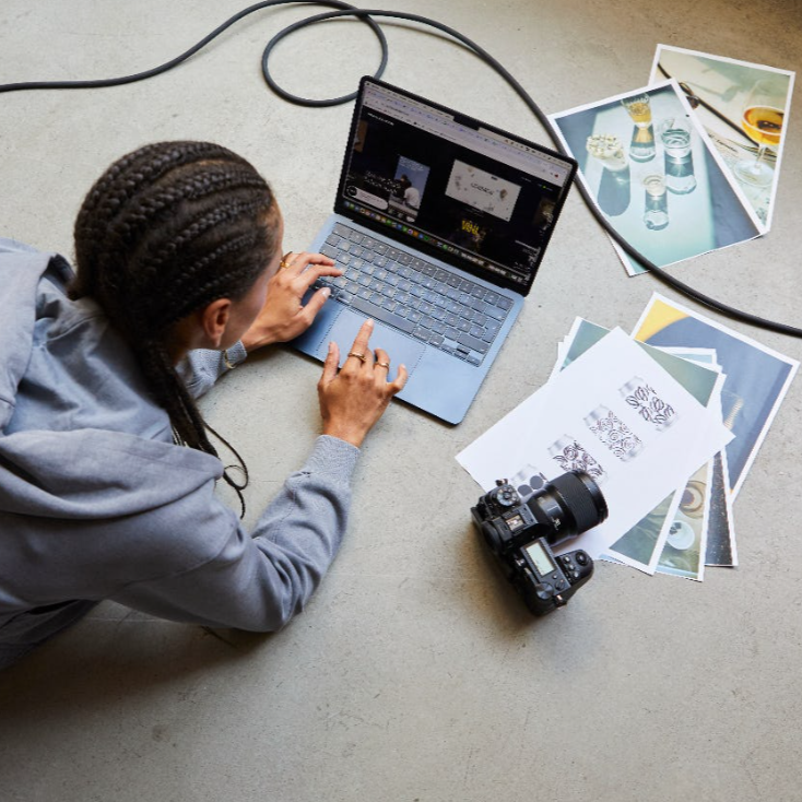 A person with braided hair working on a laptop, surrounded by photos, papers, and a camera on the floor.