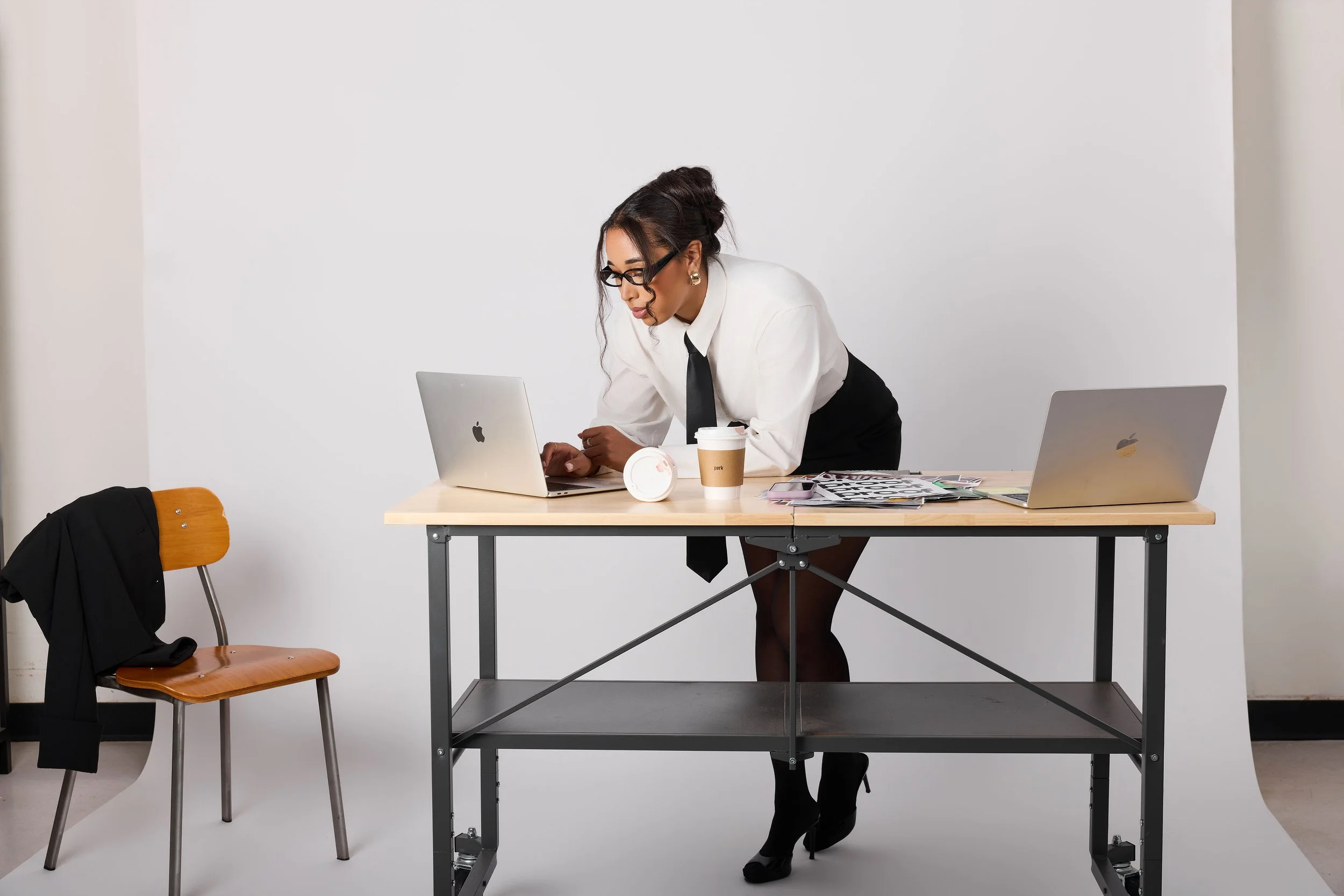 A woman with glasses, wearing a white shirt and black tie, leaning over a table with two laptops, a coffee cup, and papers, in a plain white room.