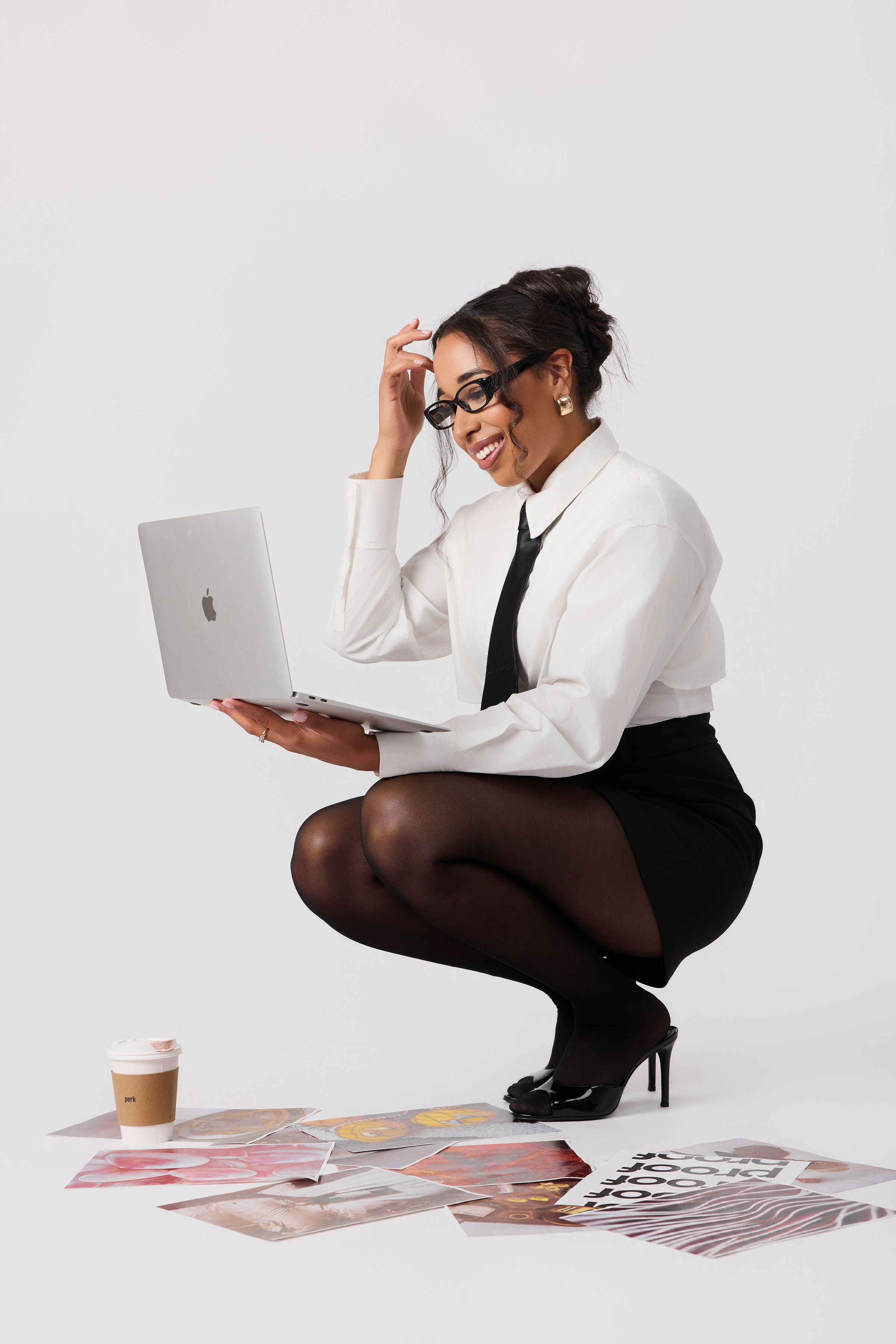 A woman dressed in professional attire, wearing glasses, black high heels, sitting on her heels with her legs tucked underneath, holding an open laptop. There are papers, magazines, and a coffee cup on the floor in front of her, against a plain white background.