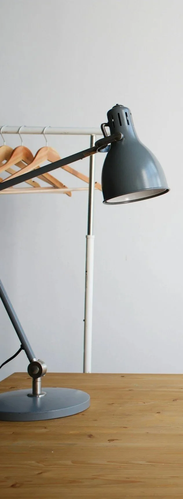Desk lamp on a wooden surface with a clothing rack and wooden hangers in the background against a plain wall.