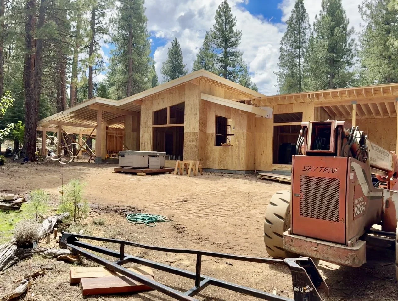 A house under construction in a wooded area with trees in the background, wooden framing, and construction equipment in the foreground.