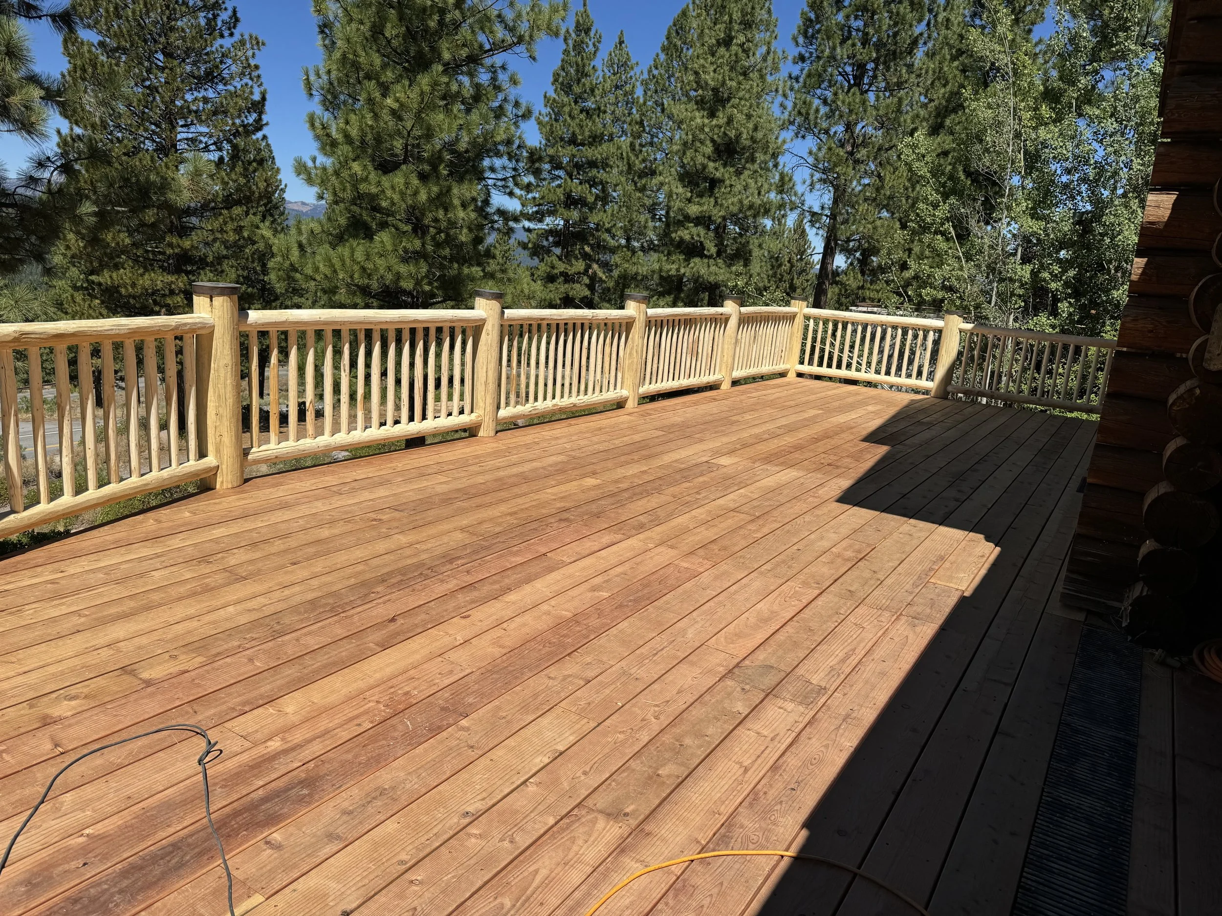 Wooden deck with railing overlooking a forested landscape with tall pine trees and a clear blue sky.
