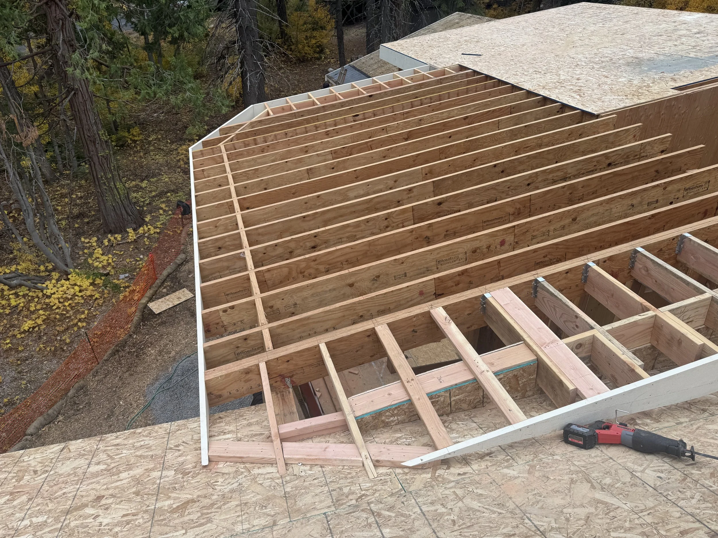 Construction site showing the framing of a new roof with wooden beams installed and some plywood sheathing partially installed, with trees in the background.