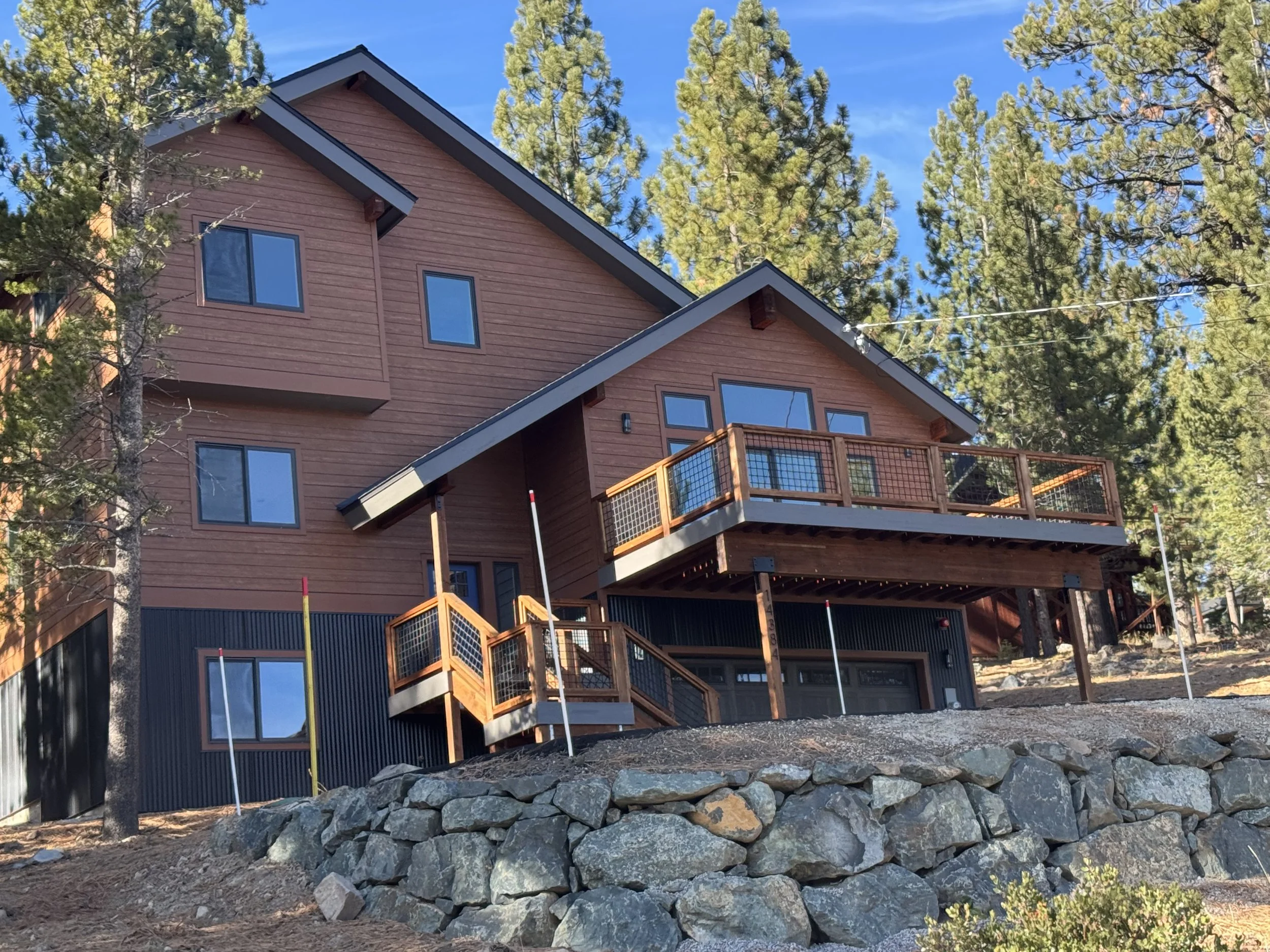Modern multi-story house with large windows, parked on a rocky terrain, surrounded by trees and blue sky.