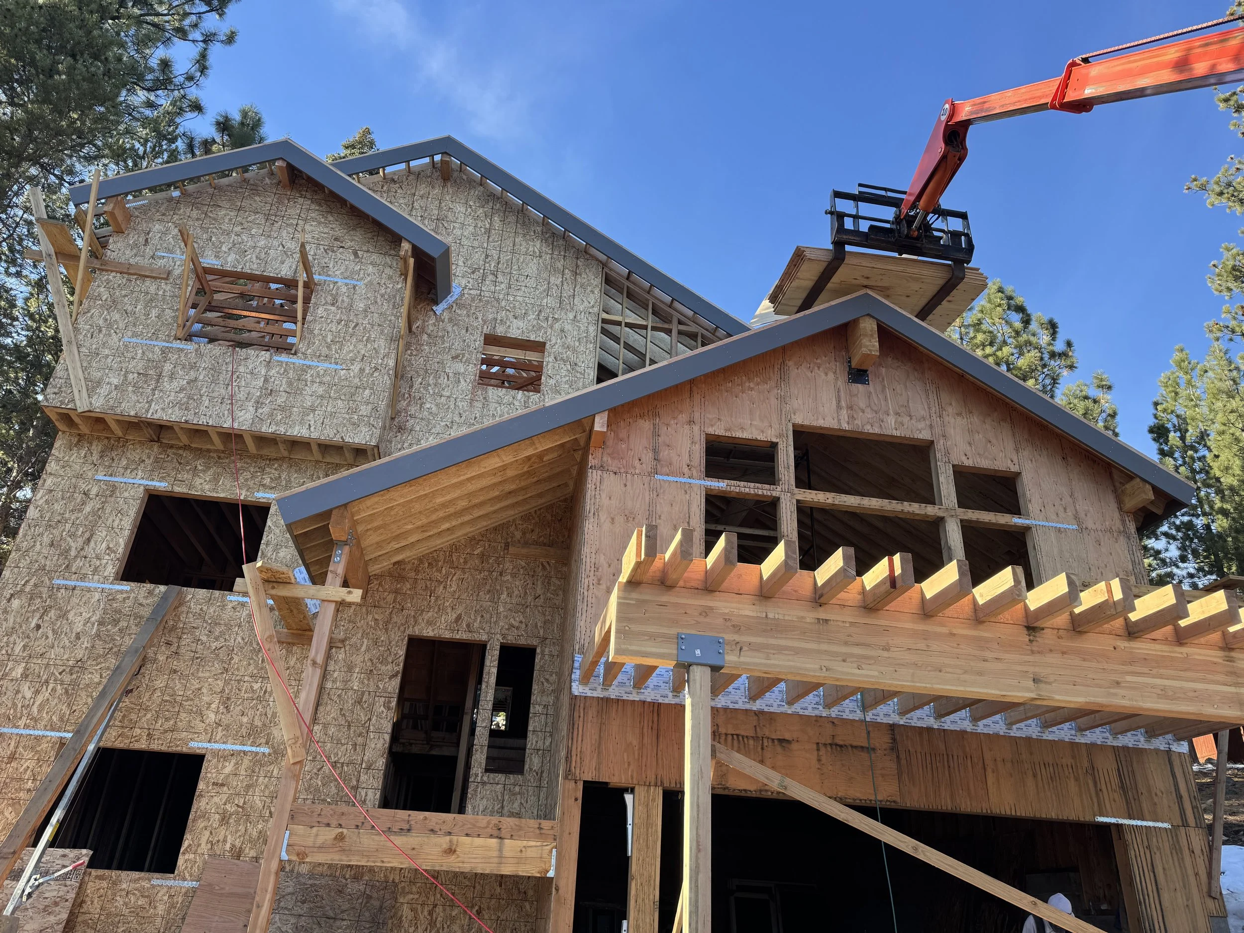 A house under construction with wooden framing, plywood sheathing, and a crane lifting a platform on the roof, surrounded by trees and a clear blue sky.