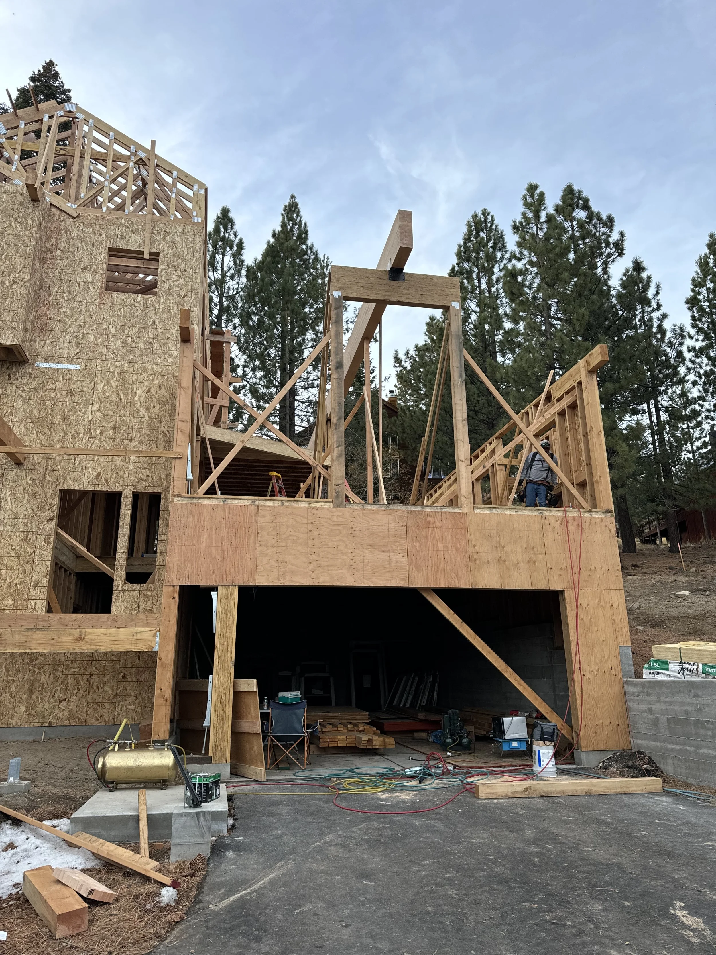 Construction site of a multi-story wooden house with framing in progress, surrounded by trees, with construction tools and materials visible.