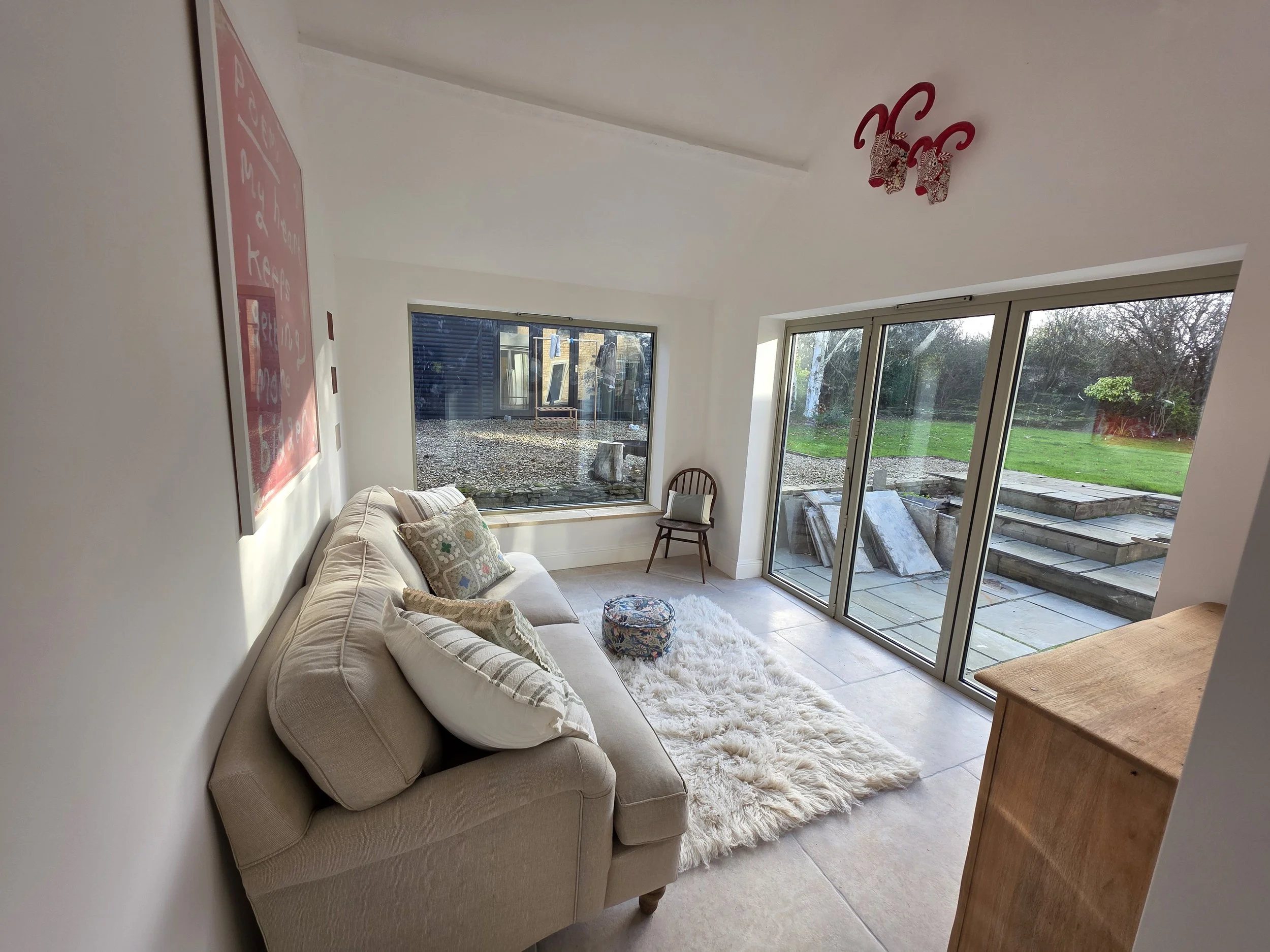 Living room with beige couch, pillows, fluffy white rug, wooden chair, large sliding glass doors leading outside, window with garden view, wooden chest, ceiling light fixture, and outdoor stone stairs.built by cortis construction