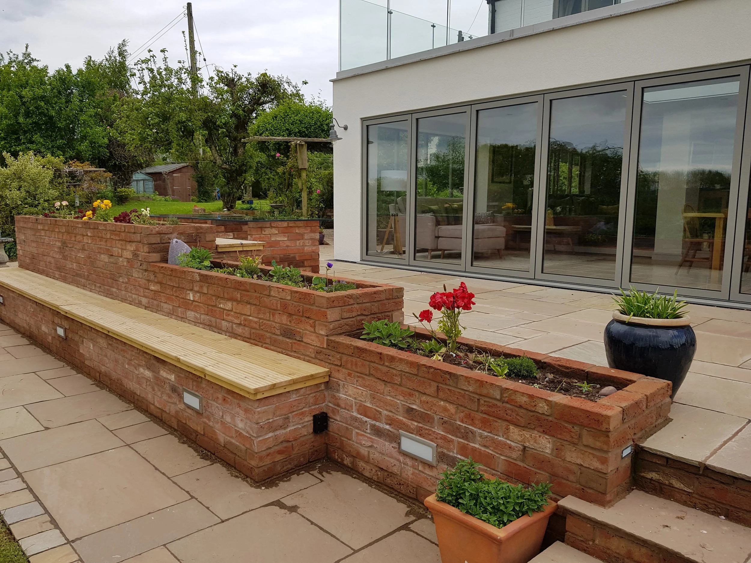Brick flower bed with blooming flowers, a wooden bench, planters, and a patio outside a house with large glass sliding doors.