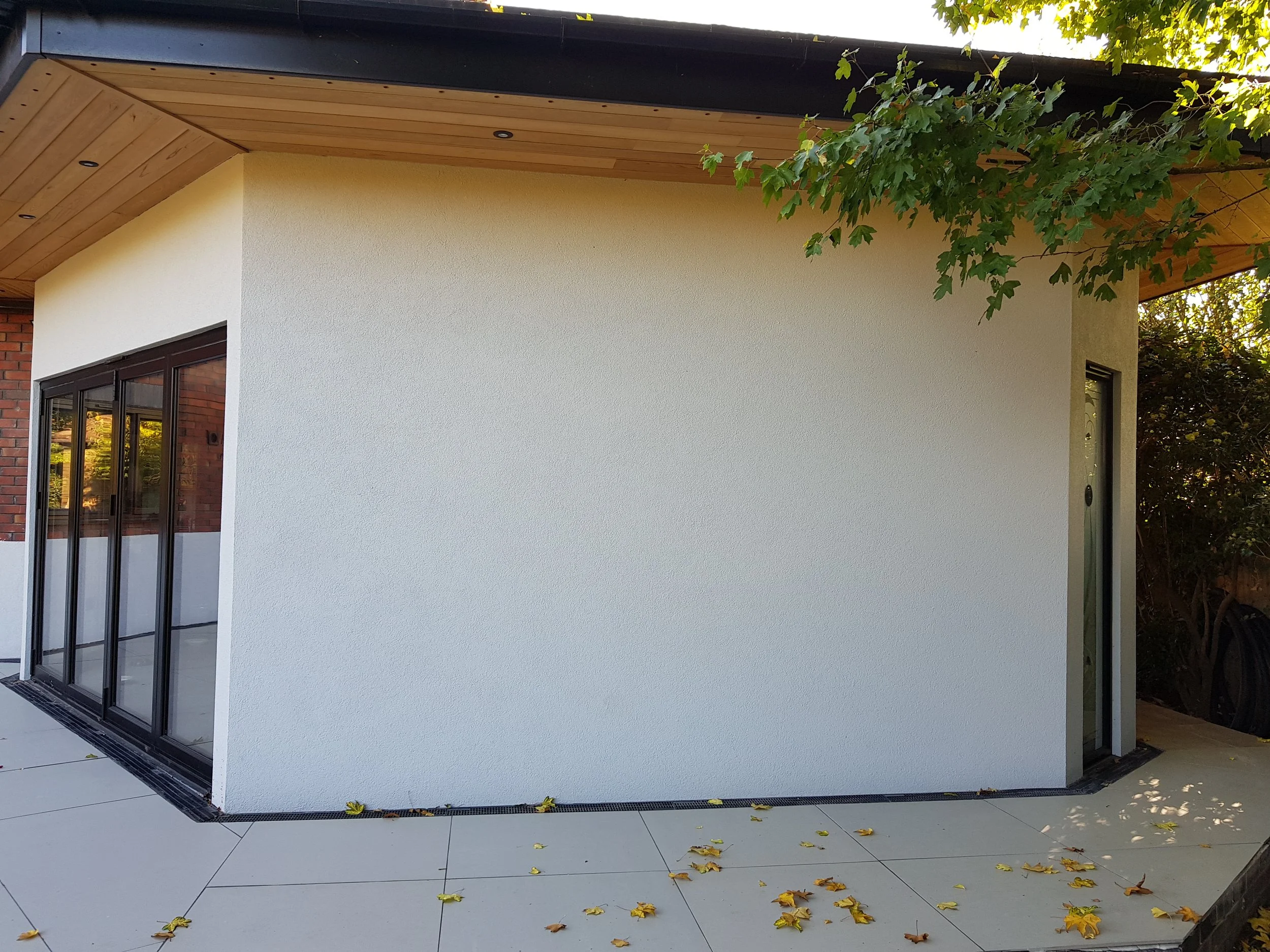 Modern corner extension with white textured exterior wall, glass sliding door, and wooden ceiling overhang, surrounded by fallen yellow leaves, on a porcelain patio laid by cortis constrcution