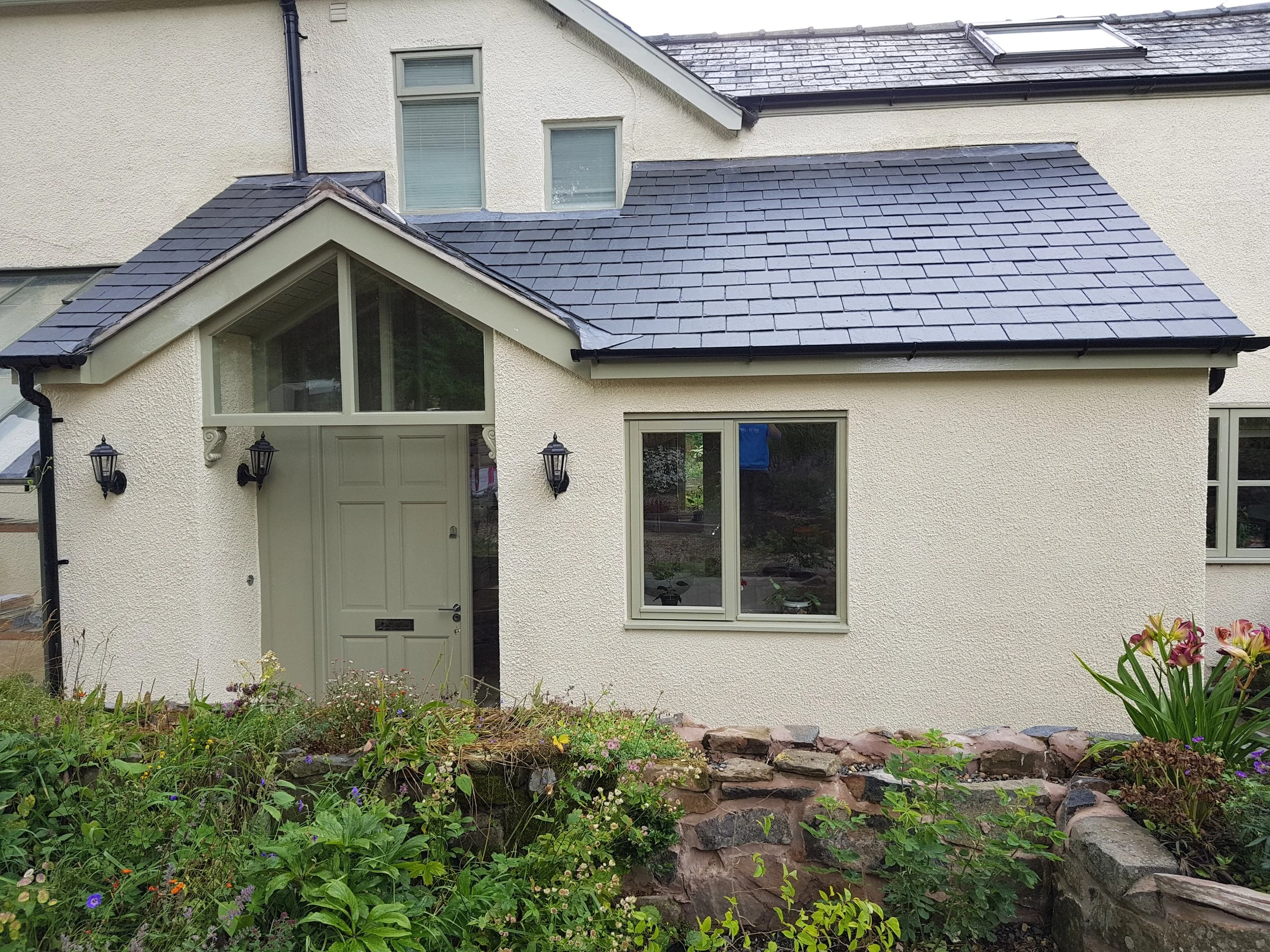 Front view of a house with light-colored stucco exterior, a pitched roof with slate shingles, and a small garden with flowers and rocks in the foreground.
