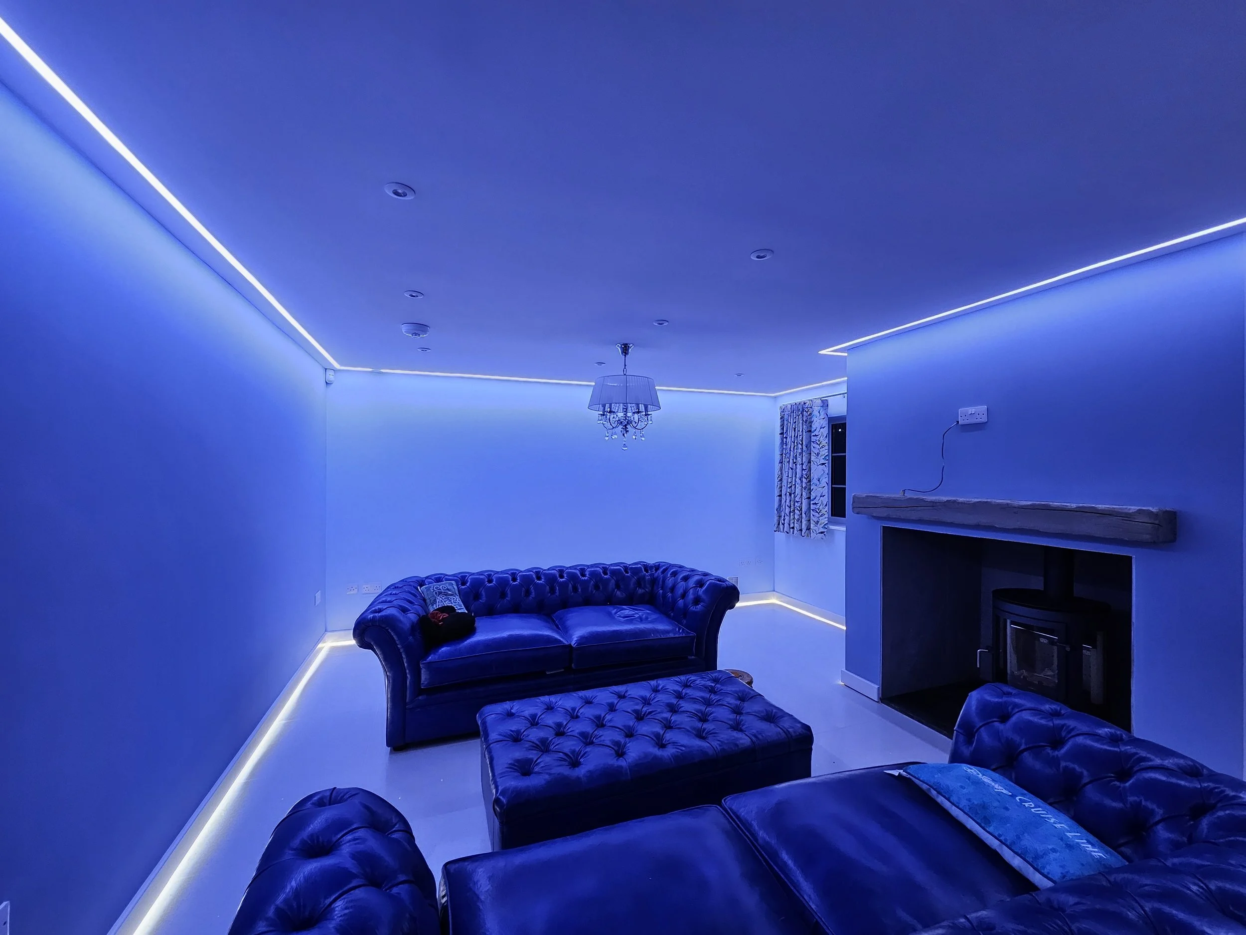 Living room illuminated with blue LED strip lights along the ceiling and baseboards, containing two dark leather tufted sofas, an upholstered ottoman, a fireplace with a mantle, a window with patterned curtains, and a chandelier hanging from the ceiling.