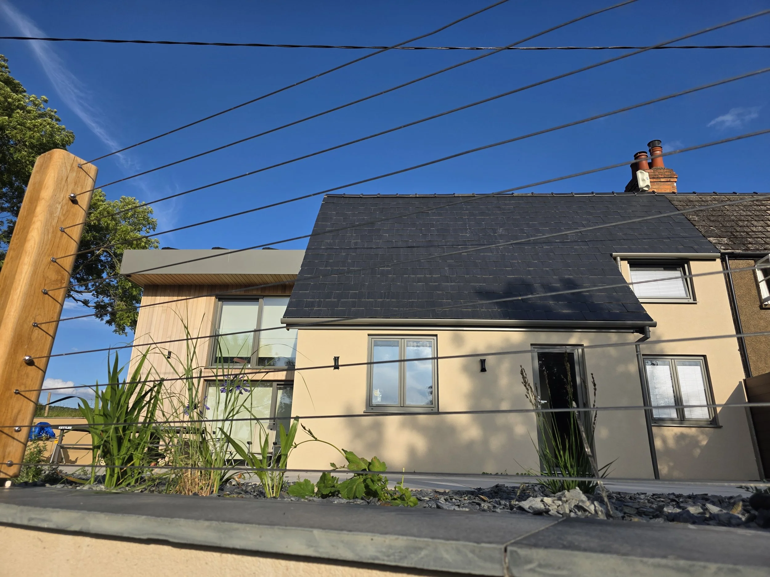 Photo of a modern house with a mix of black and beige exterior walls, large windows, and a dark tiled roof, set against a bright blue sky.