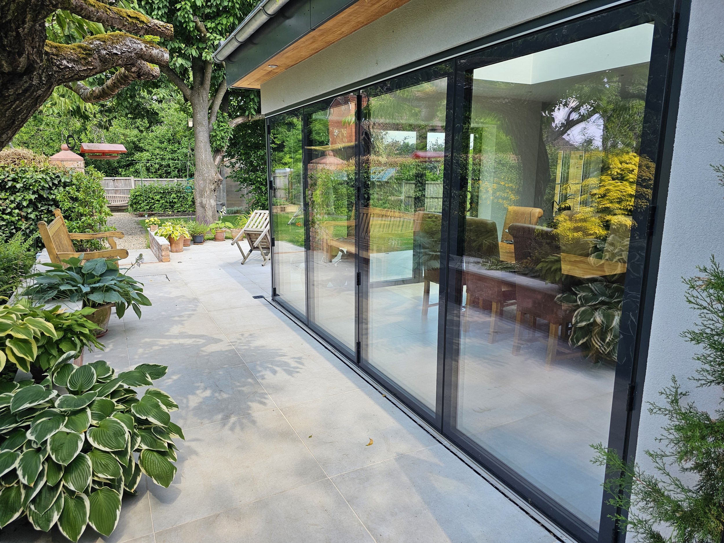 A modern house with large sliding glass doors, a concrete patio, and surrounding lush greenery including potted plants, bushes, and trees.