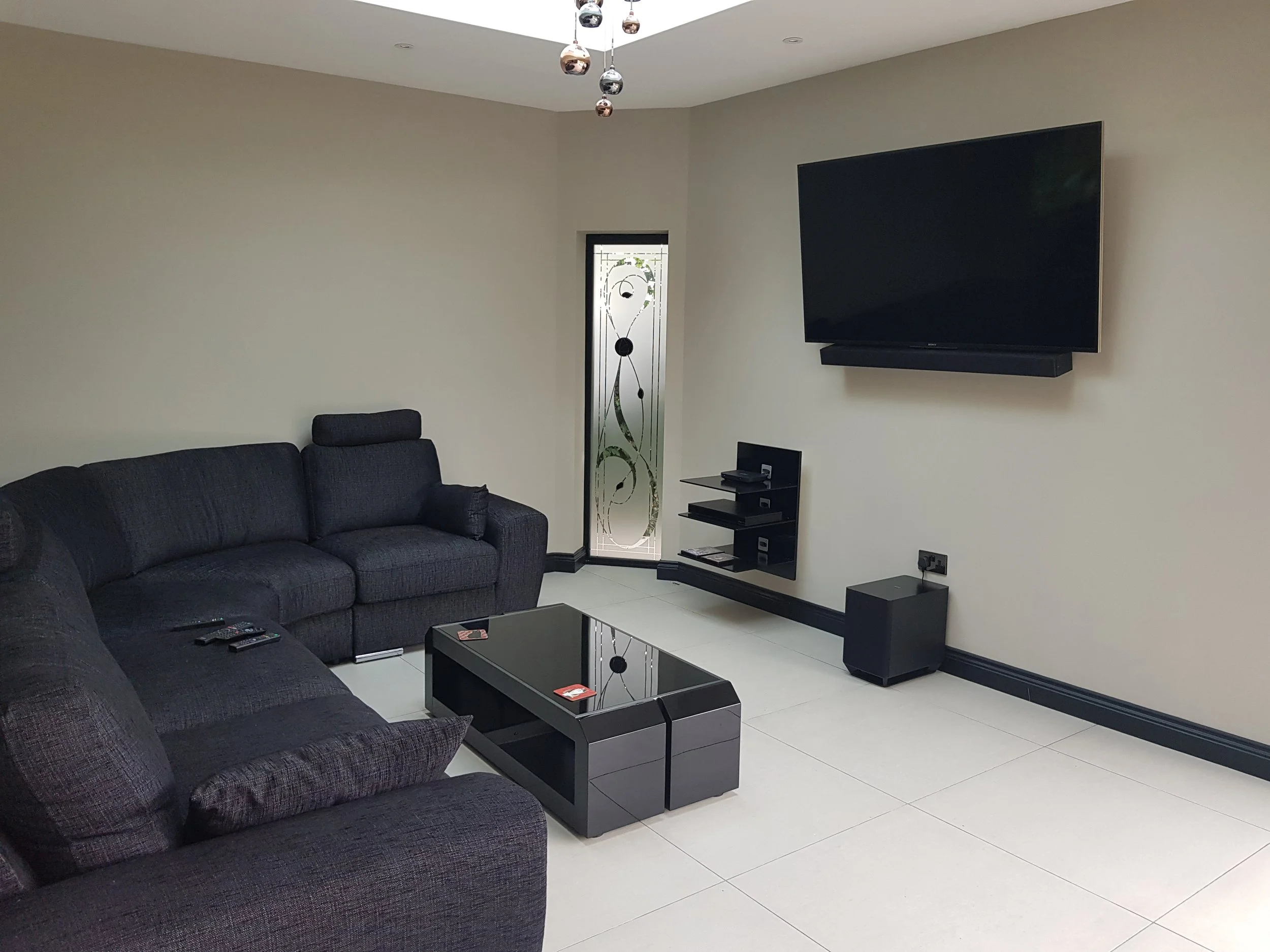 A modern living room with black fabric couches, a black glossy coffee table, a large mounted flat-screen TV, a small black shelf with electronic devices, a window with decorative frosted glass, and minimalist decor.