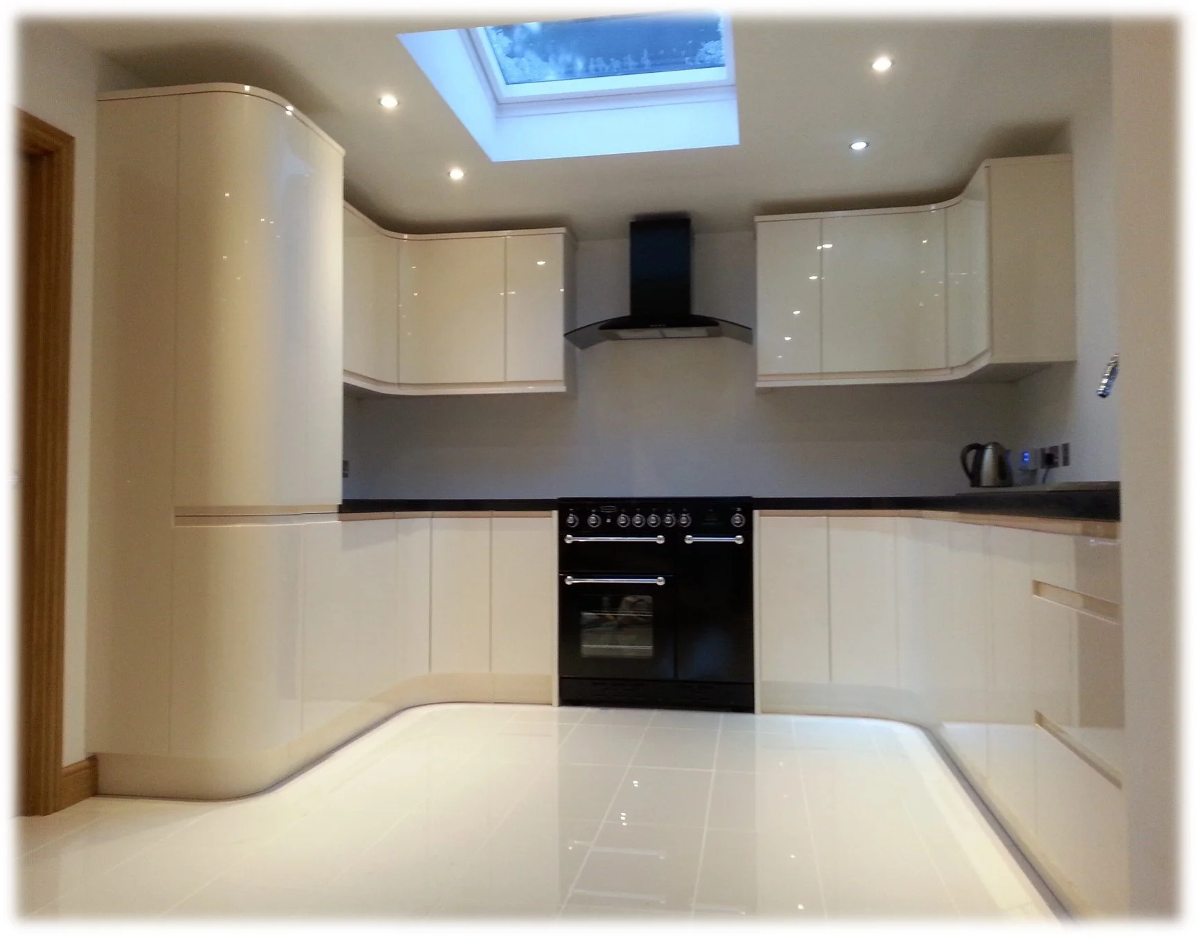 Modern kitchen with cream-colored cabinets, black oven and stove, black range hood, skylight, tiled floor, and small appliances on the counter.