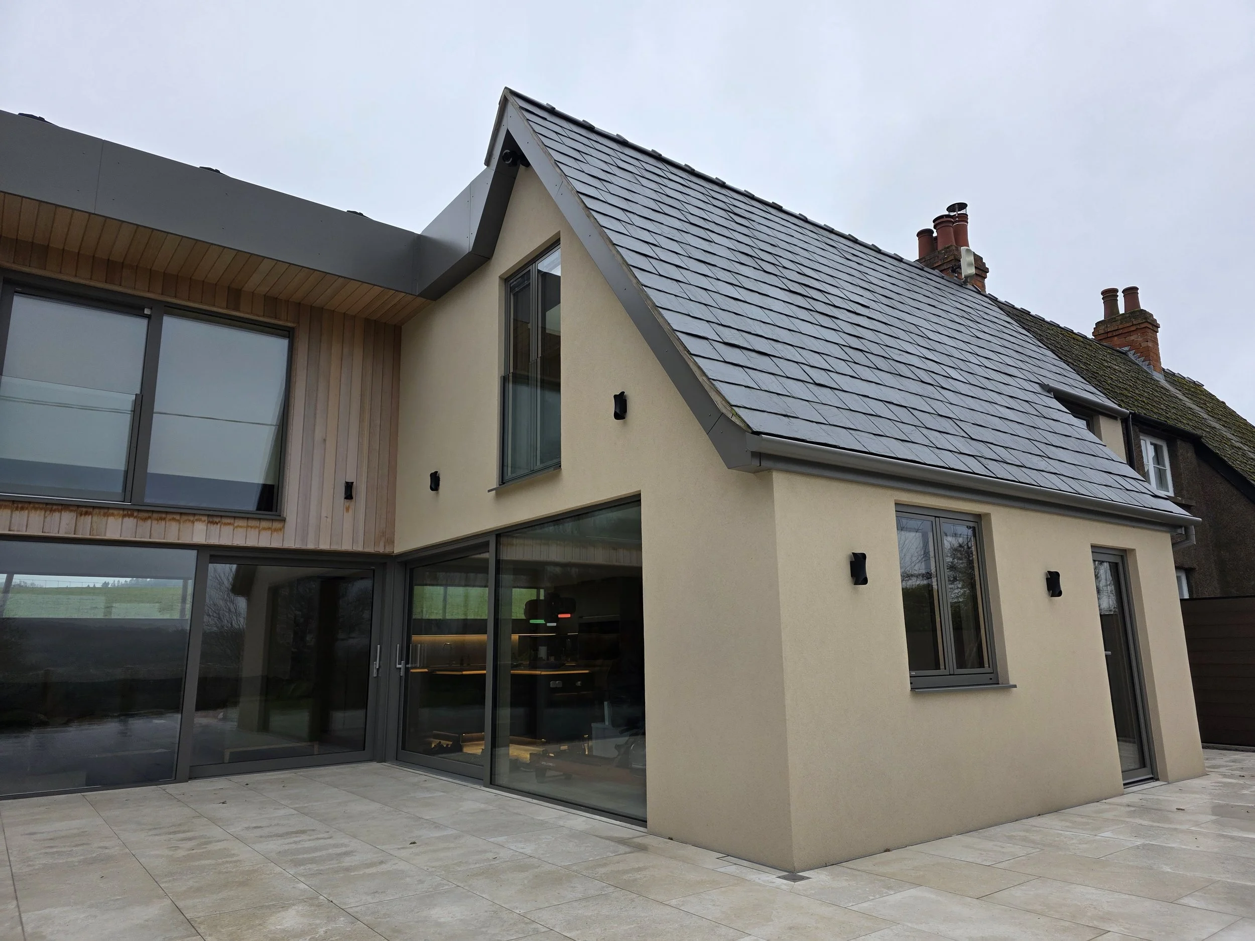 Modern house with beige exterior walls, large windows, a section with wood siding, and a gray pitched roof with chimney stacks, set against an overcast sky.
