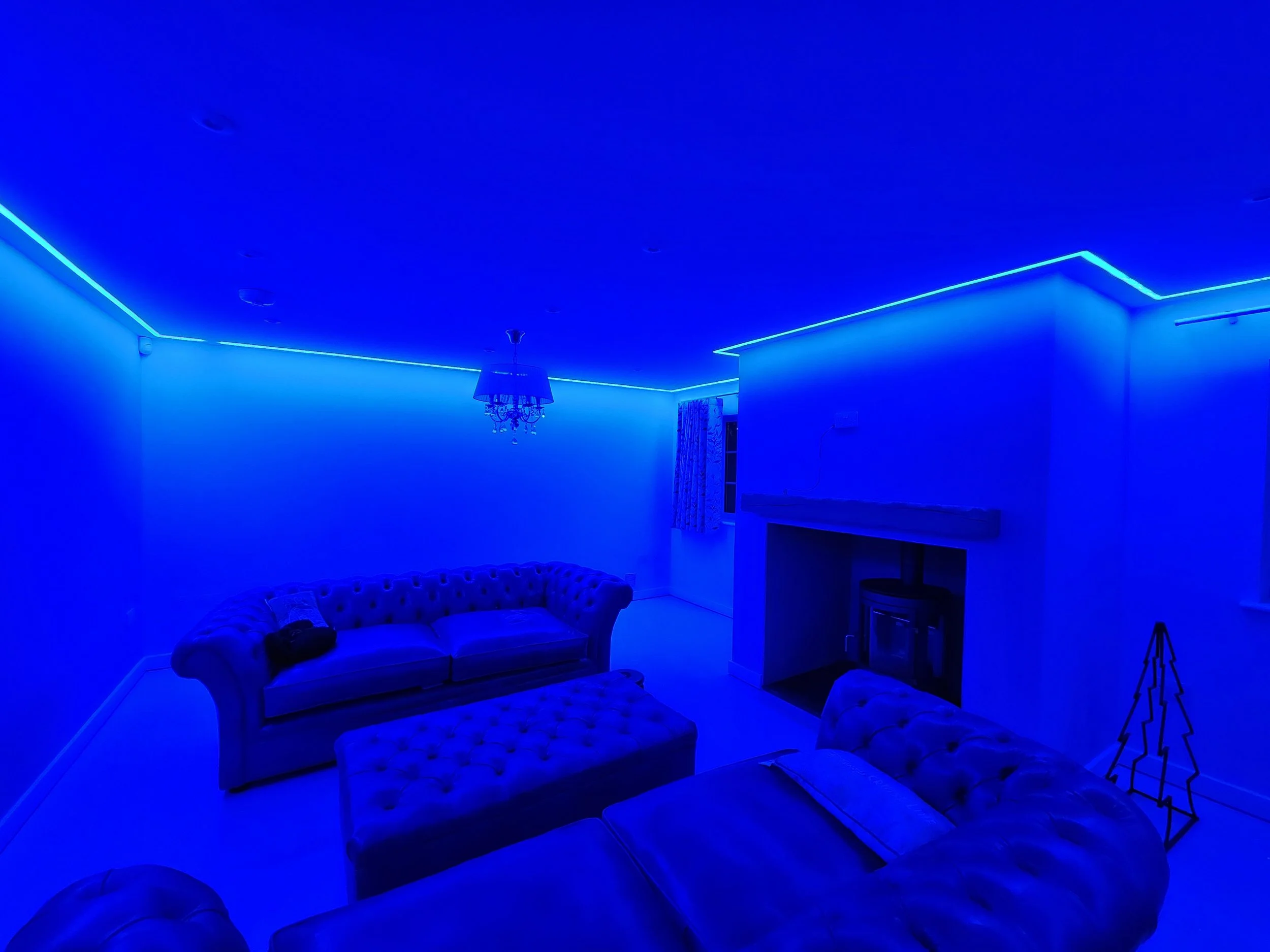 Living room illuminated with blue LED strip lights on ceiling and walls, featuring a cream-colored tufted sofa, a matching ottoman, a fireplace, a curtain window, a chandelier, and a simple metal Christmas tree.