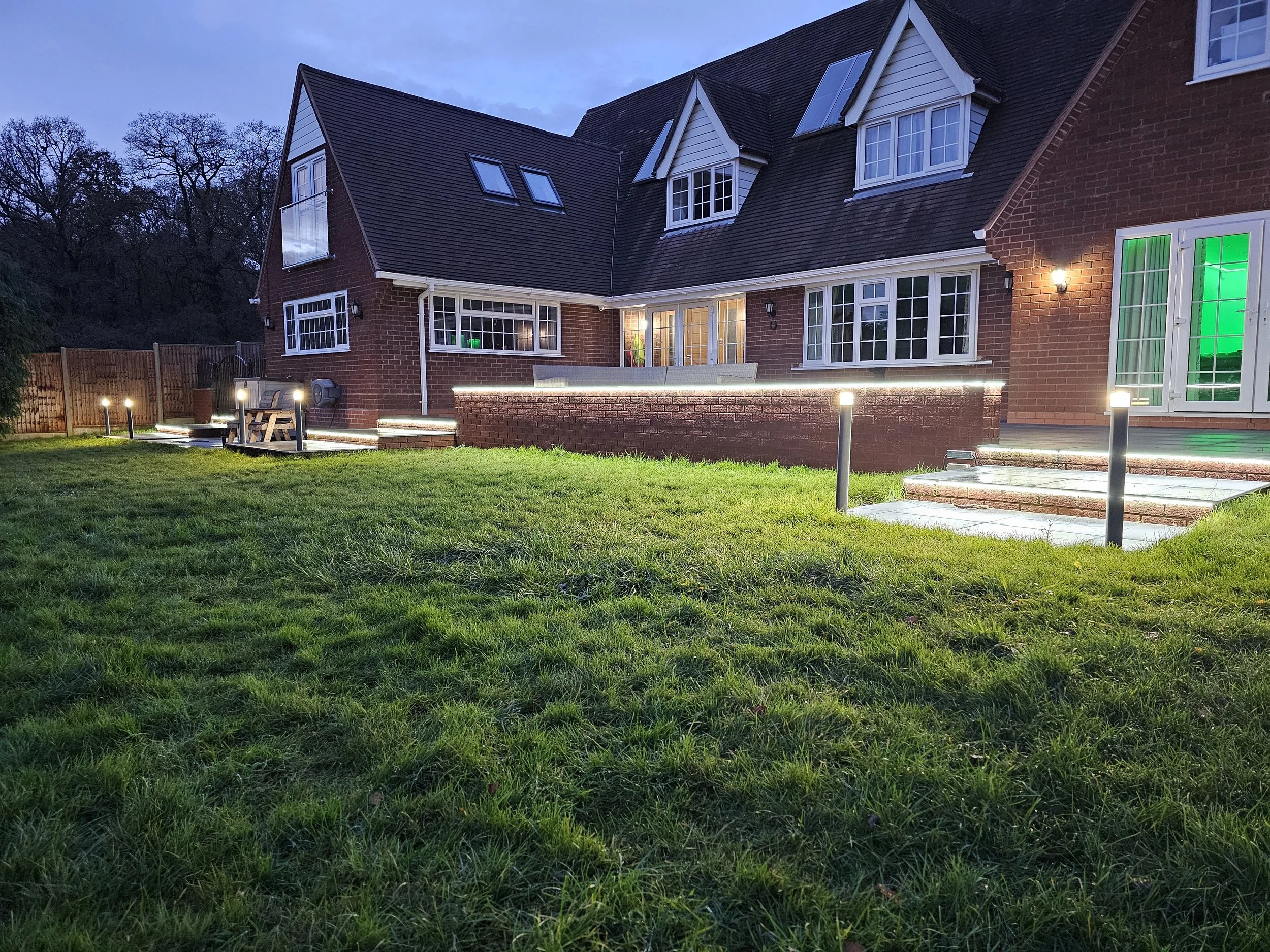 Backyard of a large brick house illuminated at dusk, featuring steps, outdoor lights, a lawn, and a fenced area surrounded by trees.