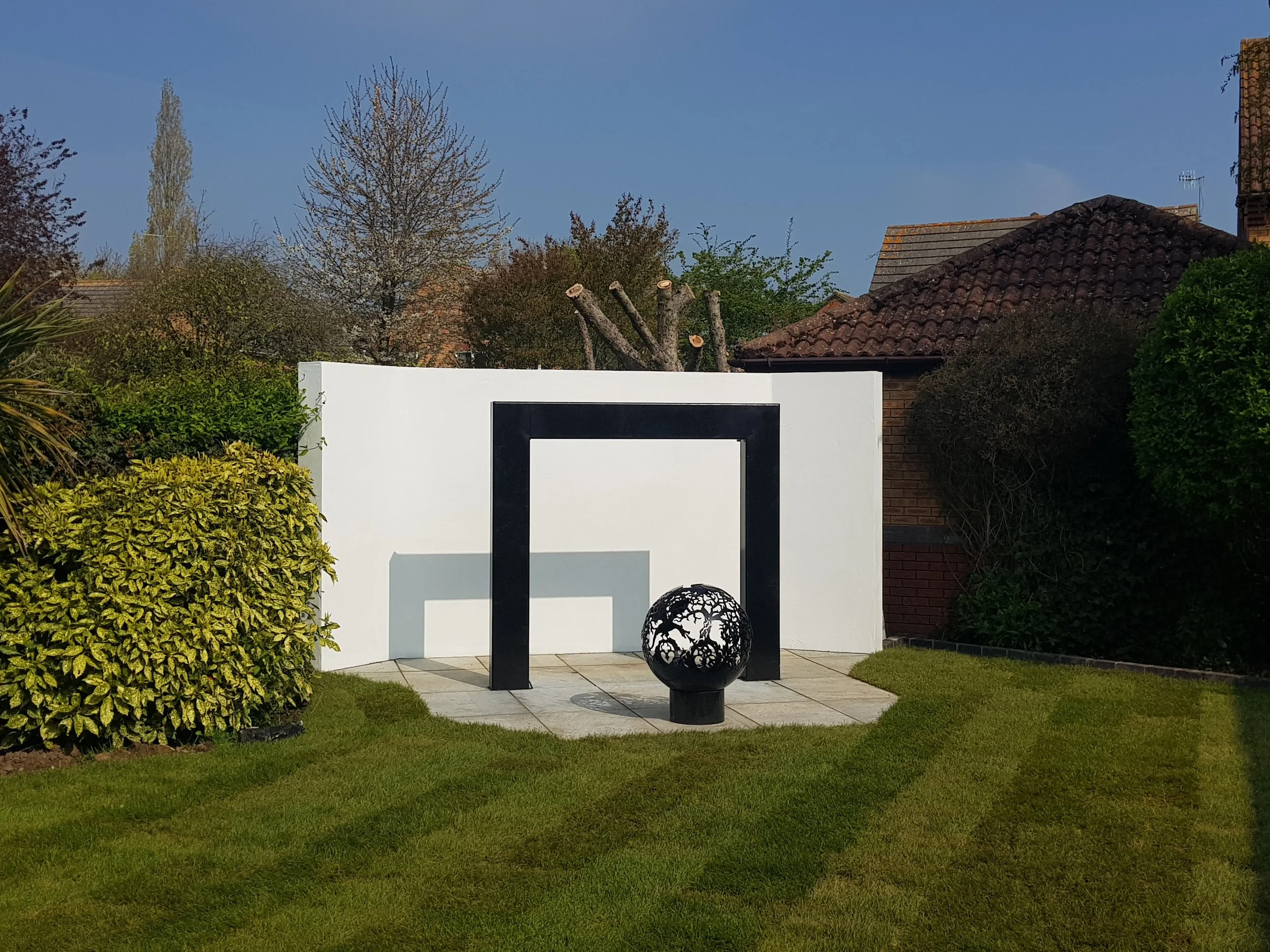 Outdoor garden with a white wall, black metal arch, spherical decorative sculpture, surrounded by greenery and neighboring houses with tiled roofs under a blue sky. all designed and built by cortis construction