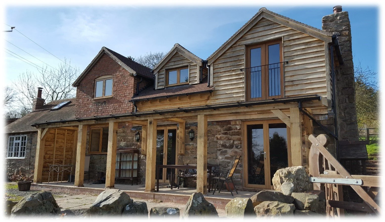 A house with a stone and wood exterior, featuring a covered patio area with wooden posts and outdoor furniture, set against a backdrop of trees and a blue sky.