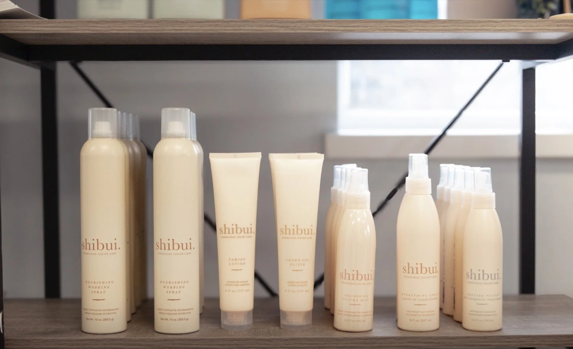 A display of beige-colored bottles and tubes of skincare products from the brand 'shibui,' arranged on a wooden shelf with a window in the background.