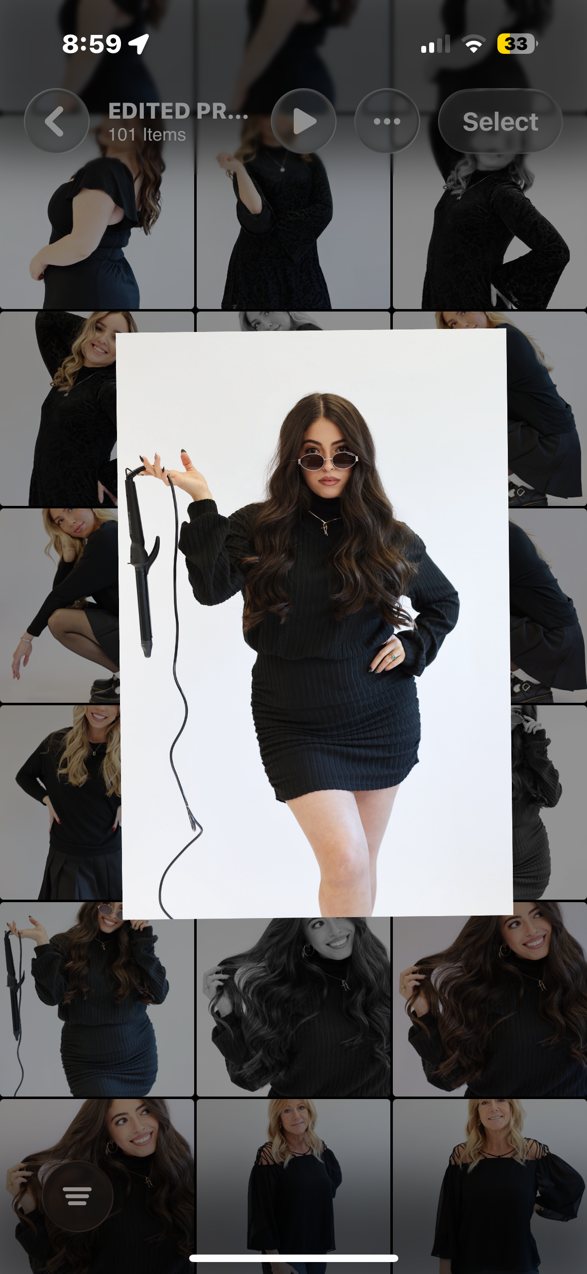 A woman with long wavy brown hair, wearing sunglasses, a black long-sleeve ribbed dress, and posing with a curling iron in her right hand against a white background.