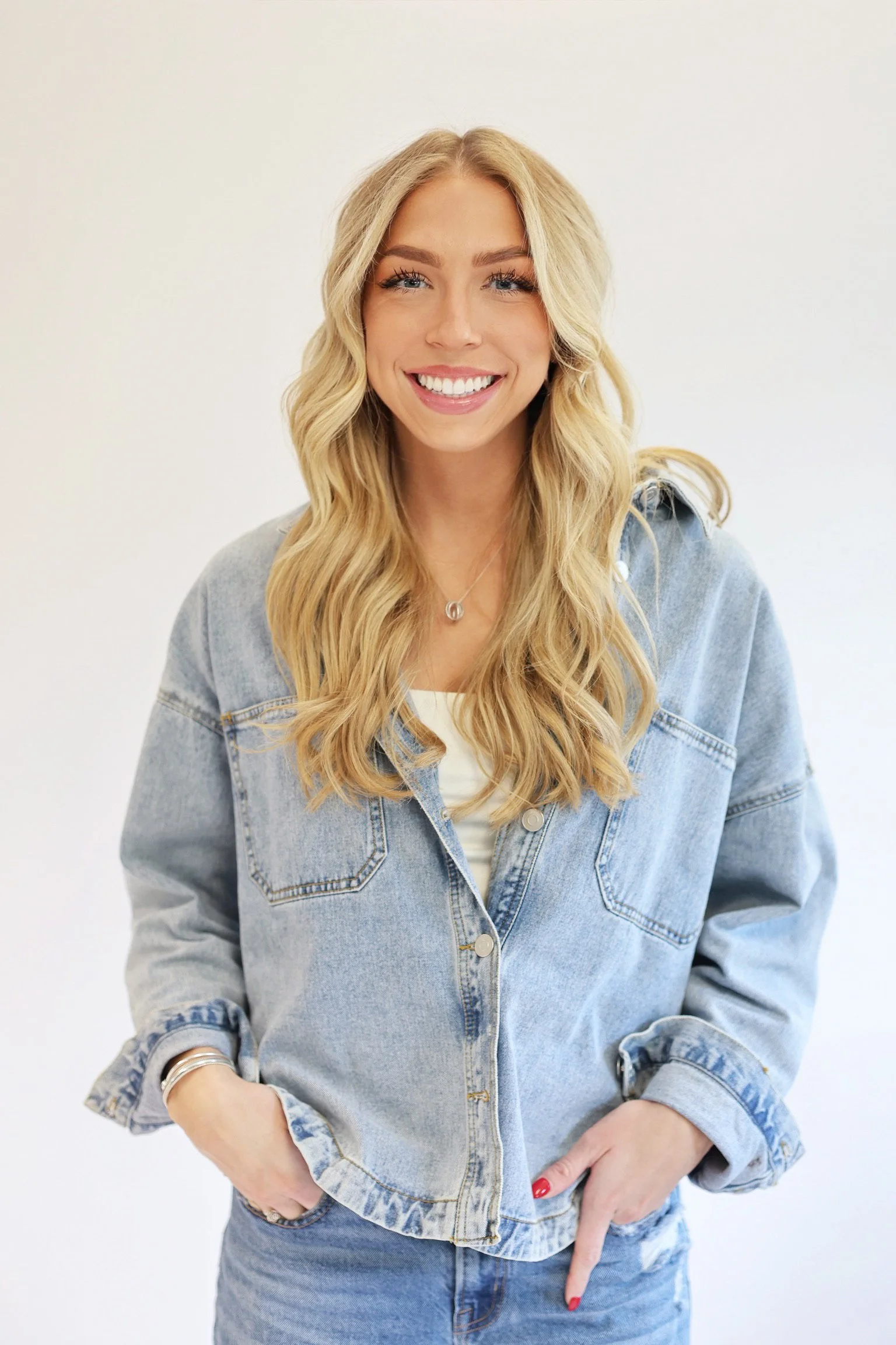 A woman with long blonde hair, wearing a light blue denim jacket and matching jeans, smiling and pointing at her waist.